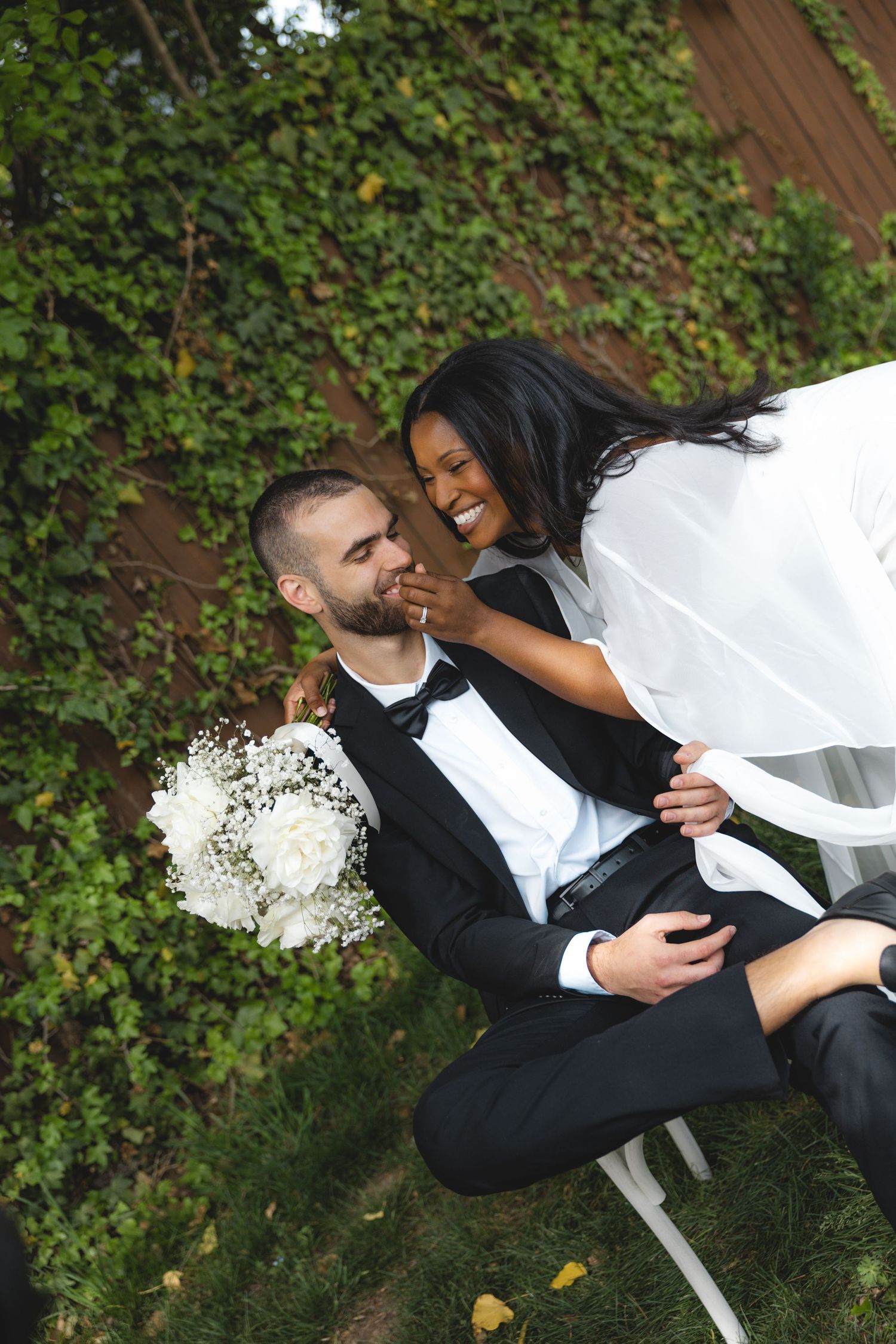 Interracial couple in wedding attire laughing outdoors, groom in tuxedo holding white bouquet, bride in veil touching his face.