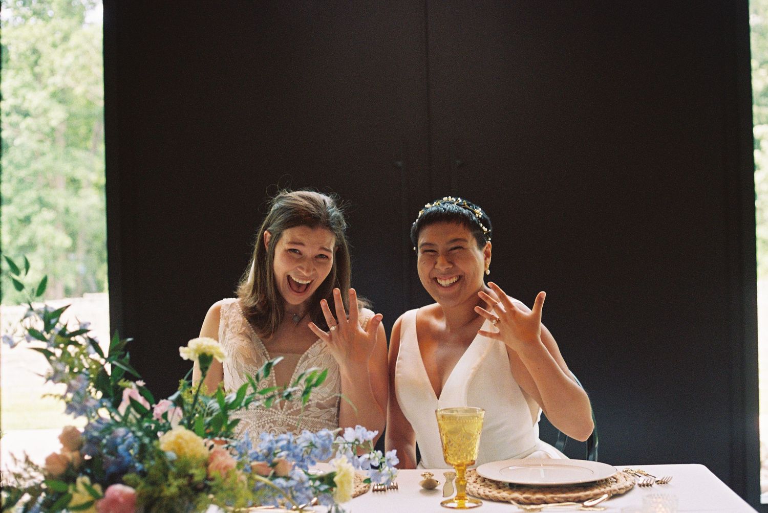 Two brides in wedding attire smile and show off rings at a floral-decorated table during their wedding reception.