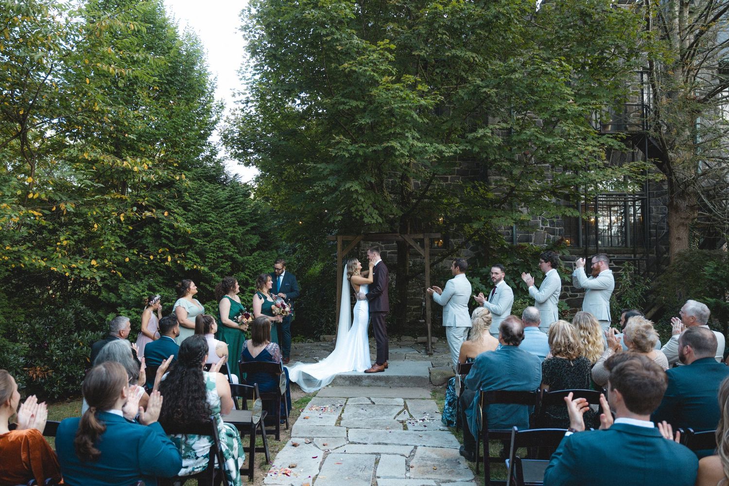 Outdoor wedding ceremony on stone patio surrounded by lush green trees, bride and groom kissing at altar with guests seated.