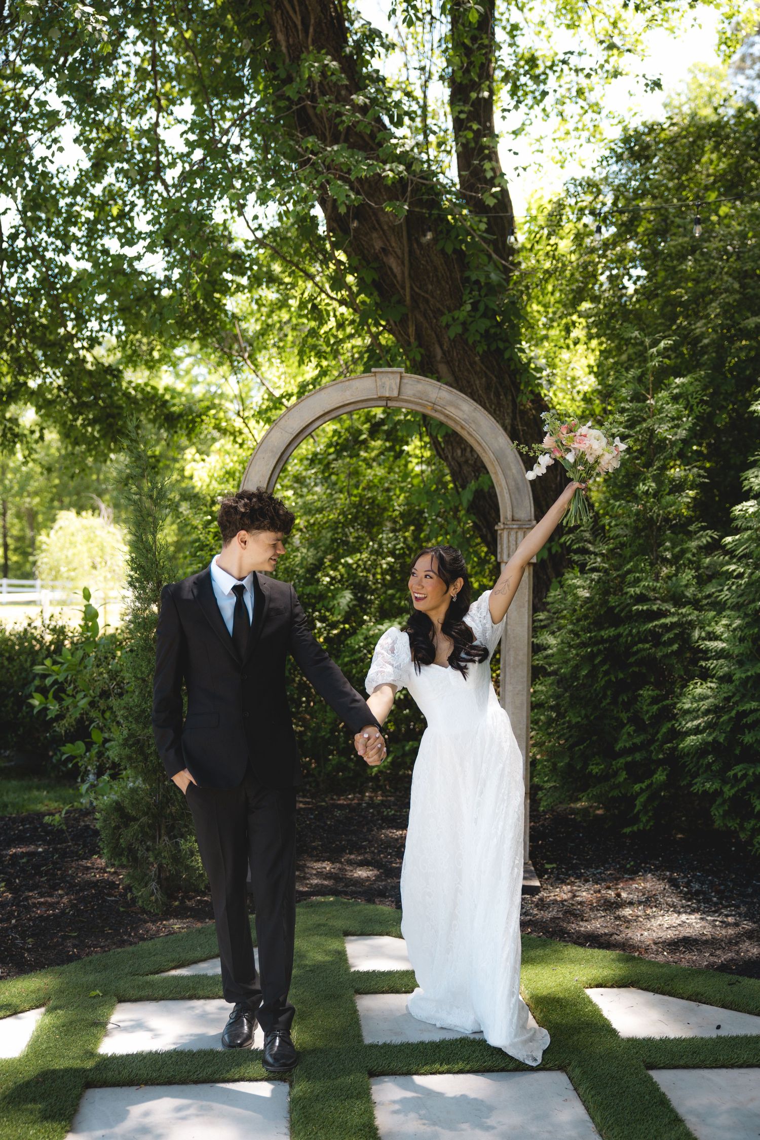 Bride in white dress raises bouquet while holding hands with groom in black suit under garden arch.