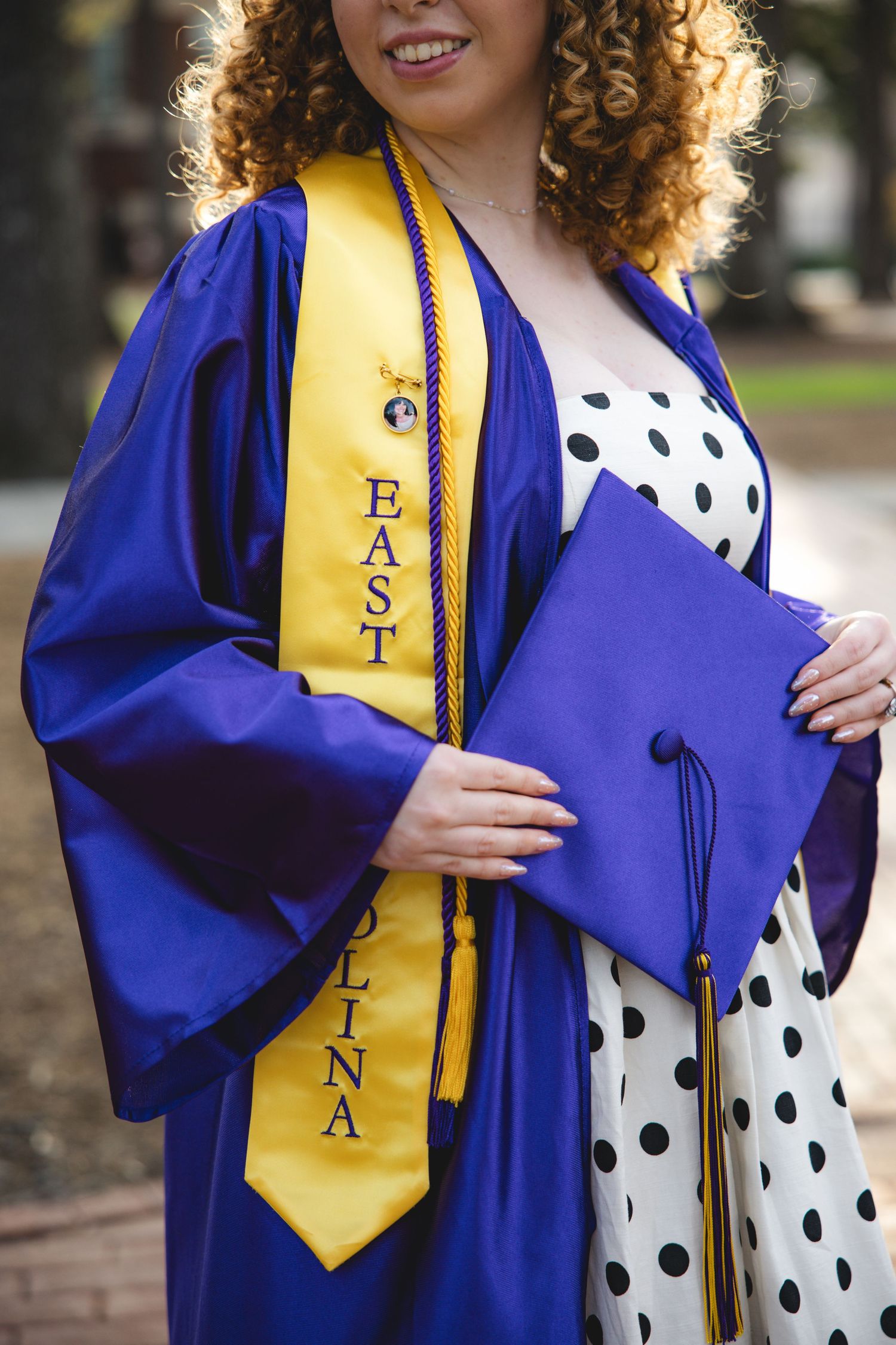 Graduate in blue and gold East Carolina University regalia holds cap, wearing polka dot dress beneath commencement gown.