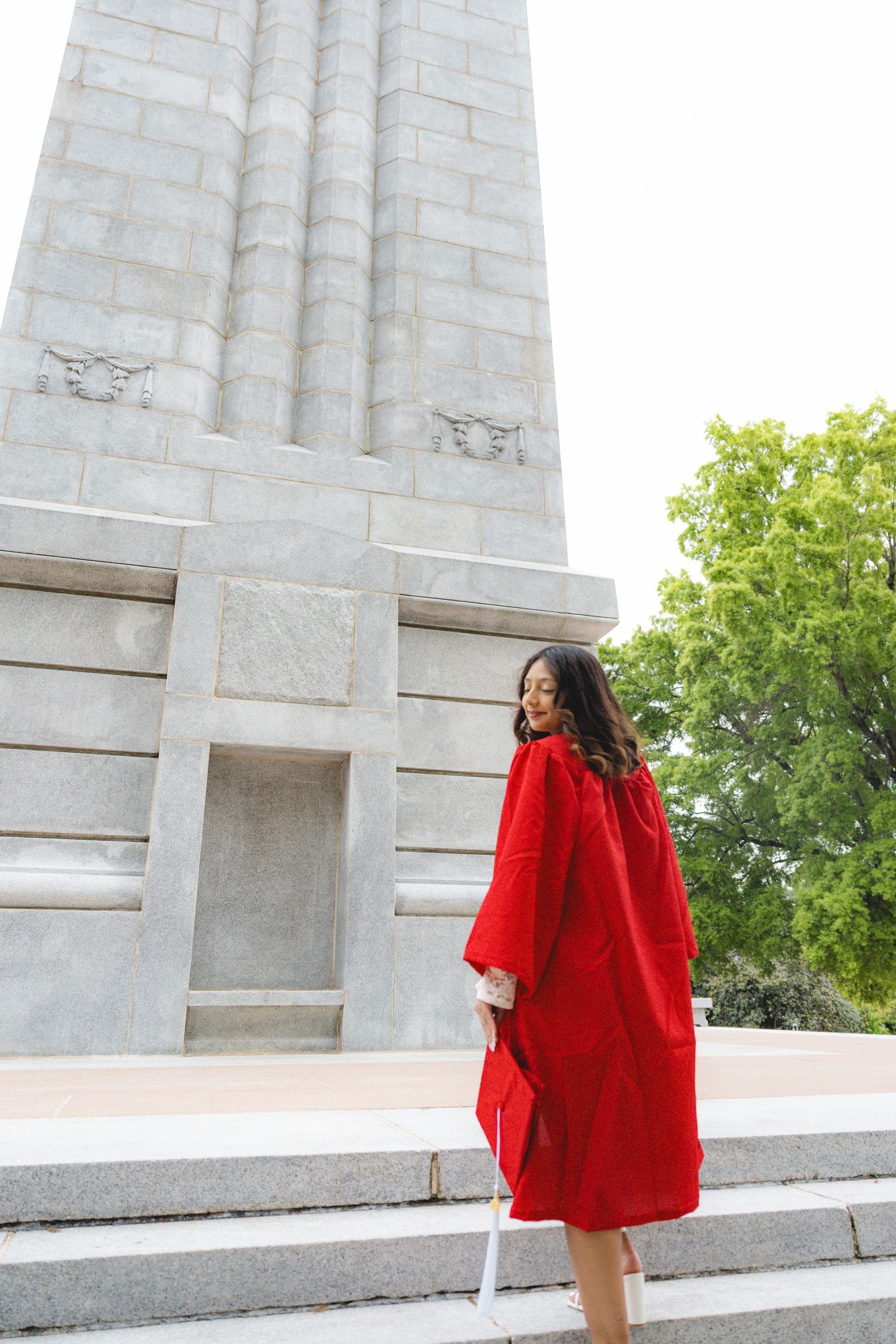 A young woman in a red graduation gown stands on steps beside a large stone monument, holding a diploma.