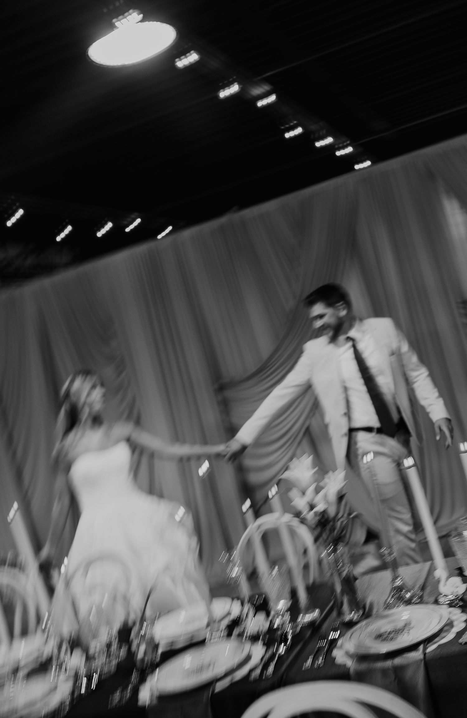 Black and white photo of bride and groom holding hands at wedding reception with elegant draped backdrop.