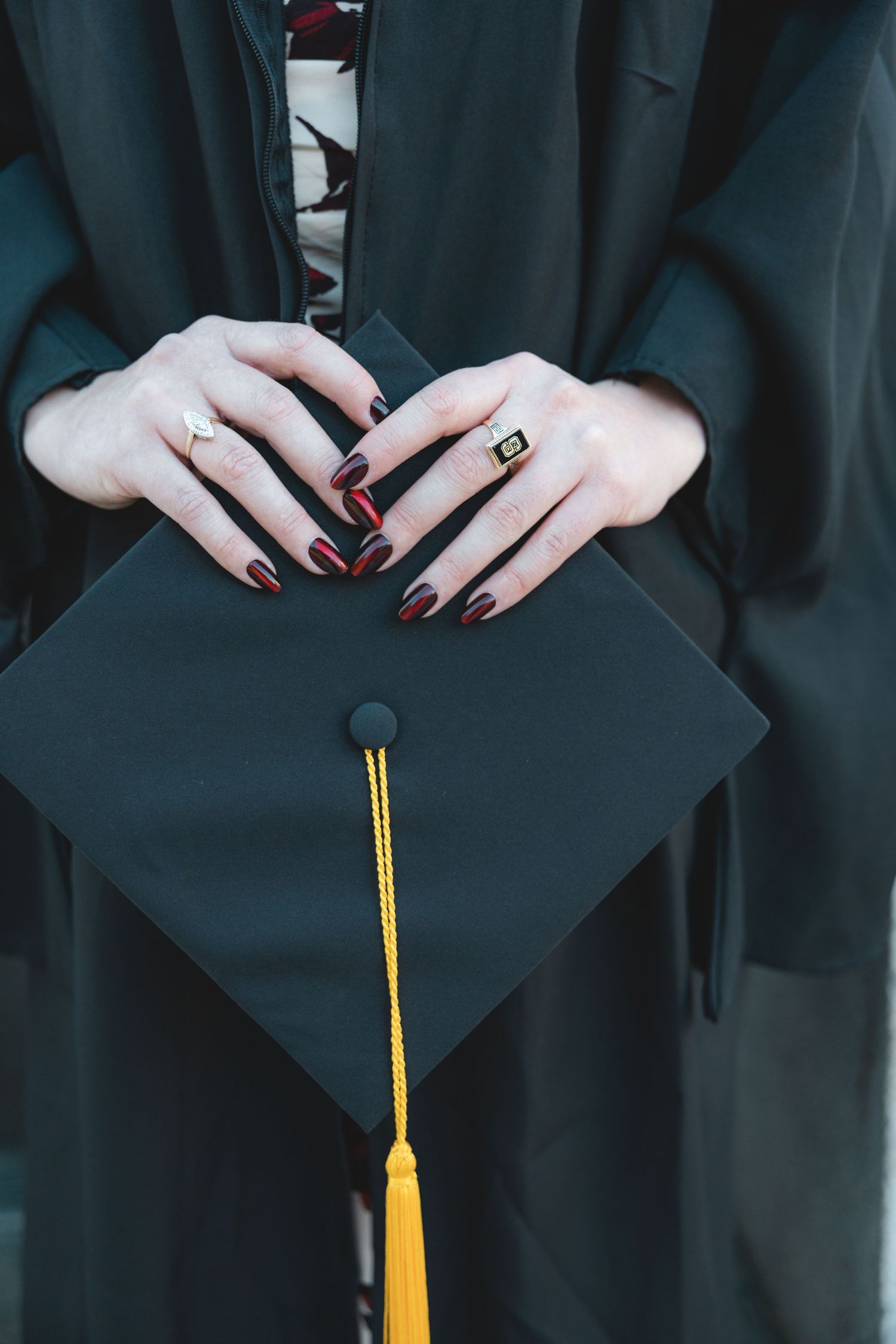 Graduate in black gown holding mortarboard cap with gold tassel, wearing rings and dark red nail polish.