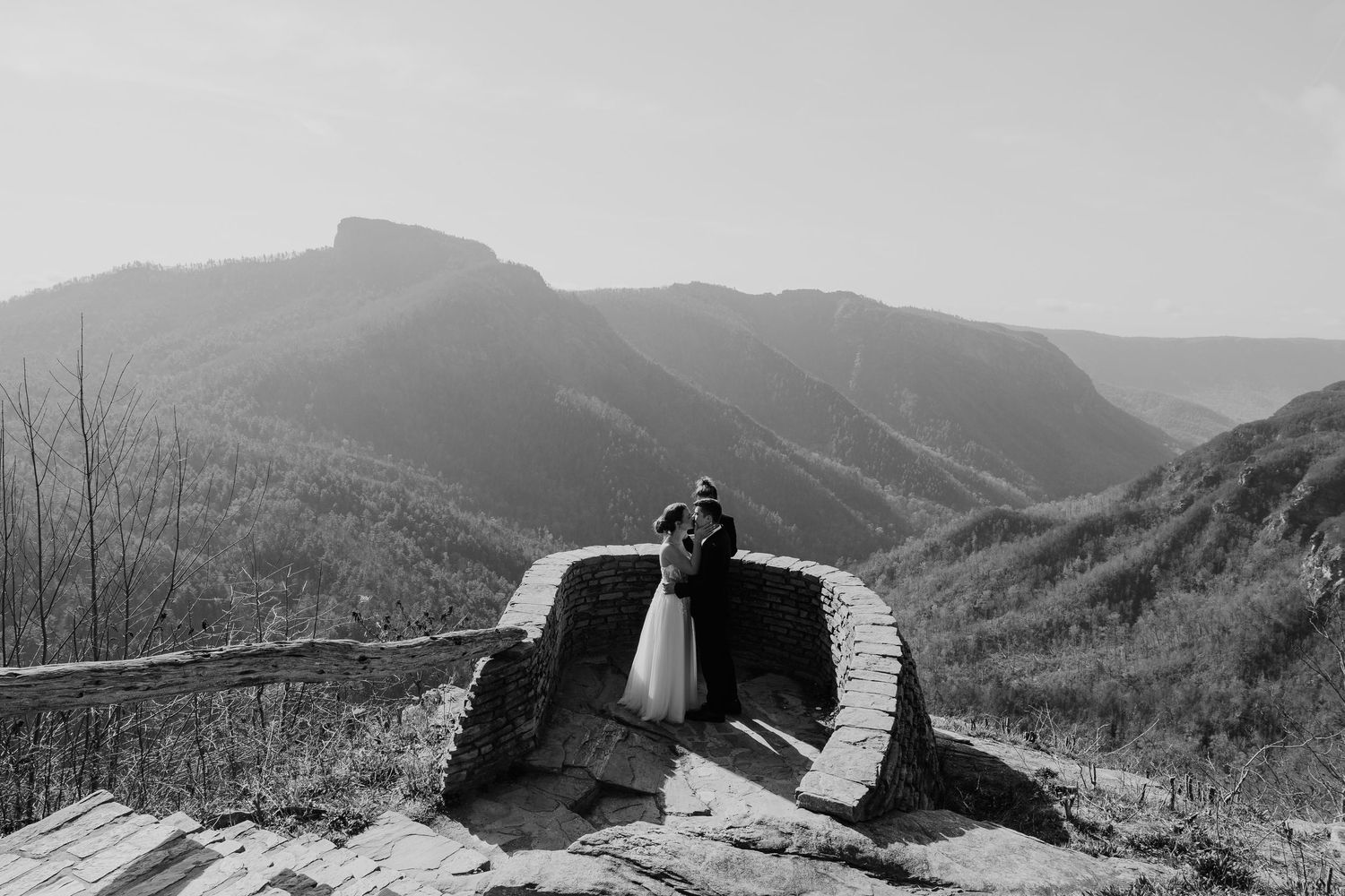 Black and white photo of a couple embracing on a mountain overlook with scenic valley and peaks behind them.