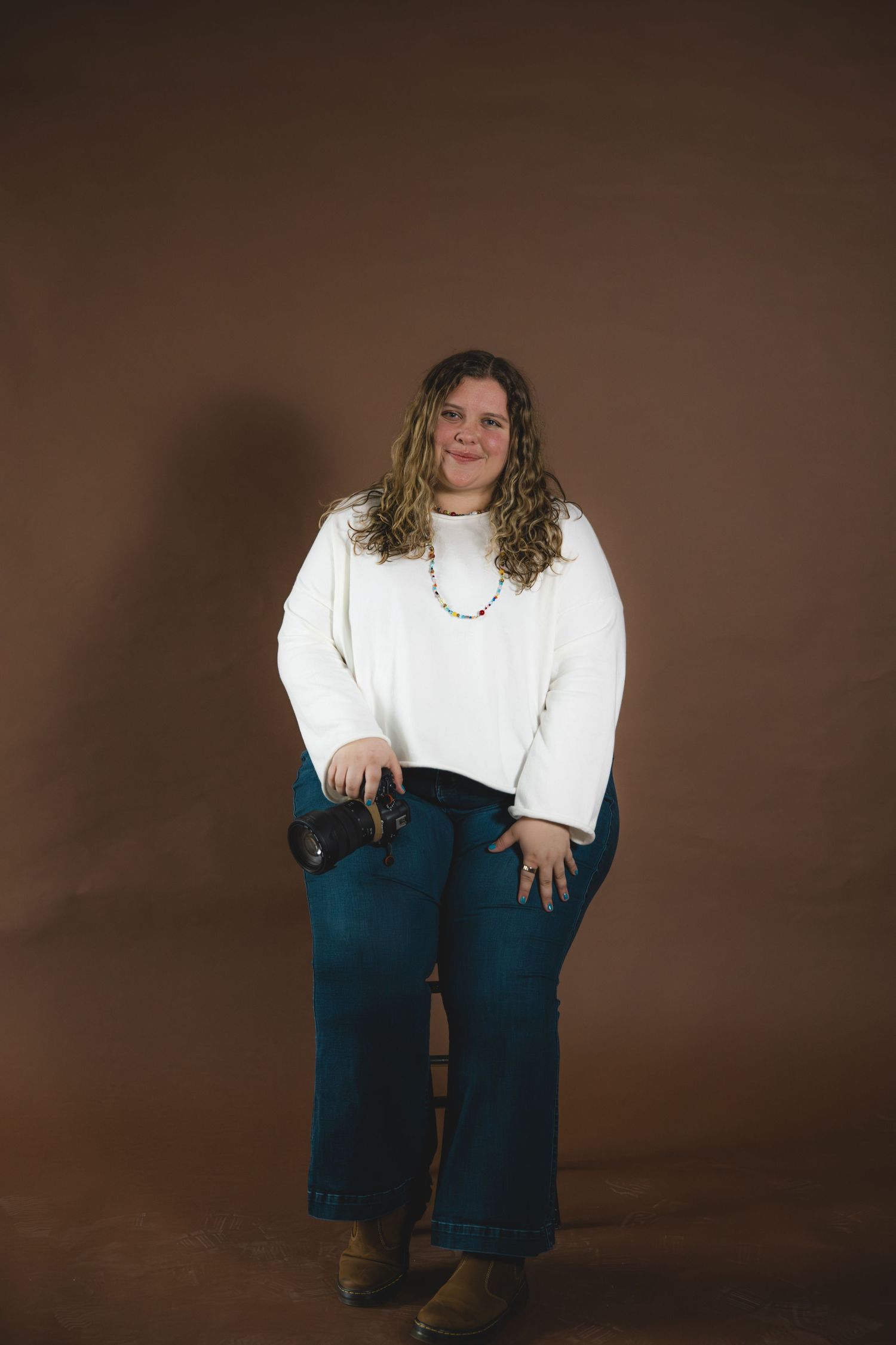 A full-length portrait of a smiling woman with curly hair wearing a white sweater against a brown background.