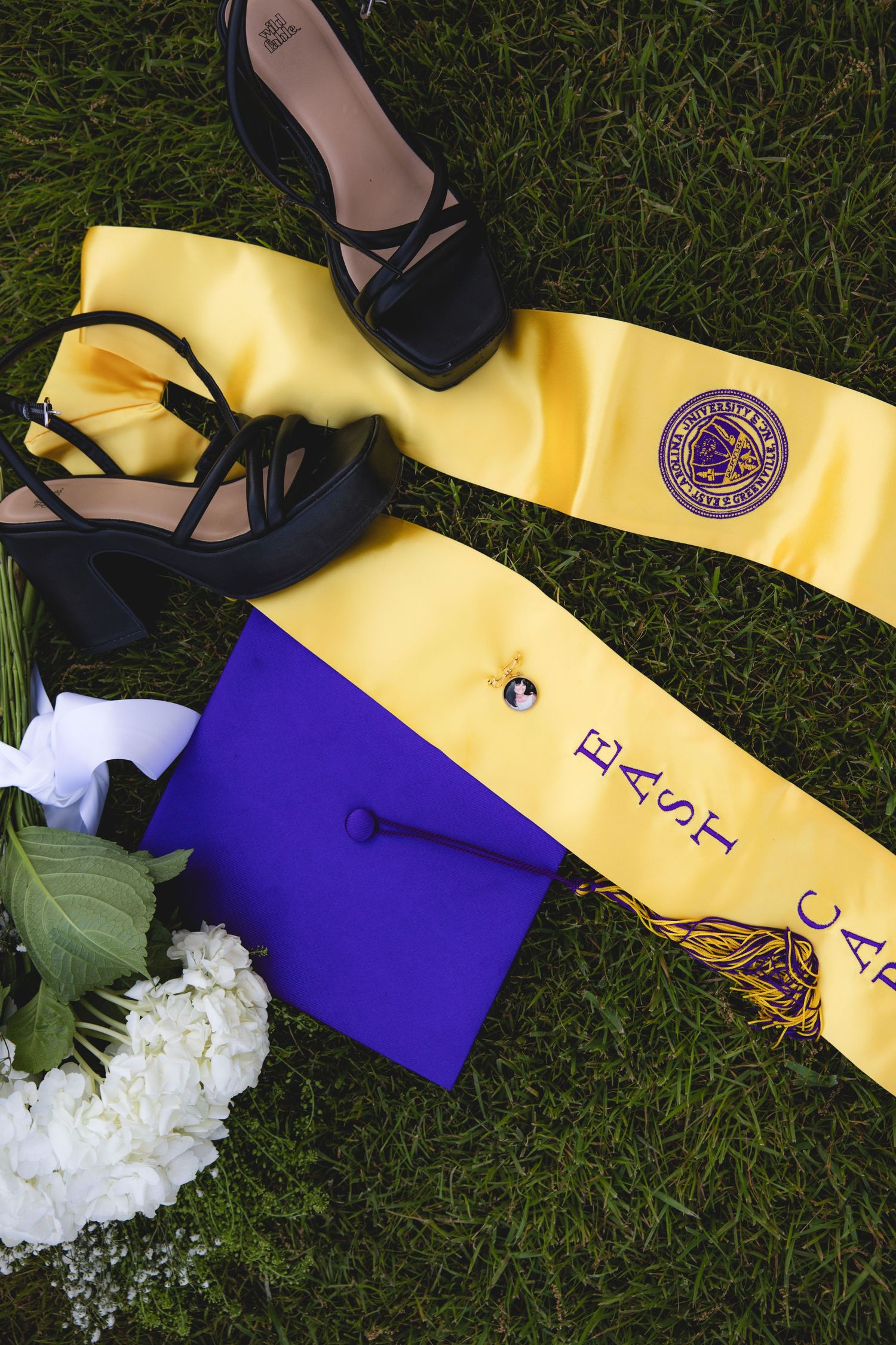 Purple graduation cap and yellow honor stole with university seal, black heels, and white flowers on green grass.