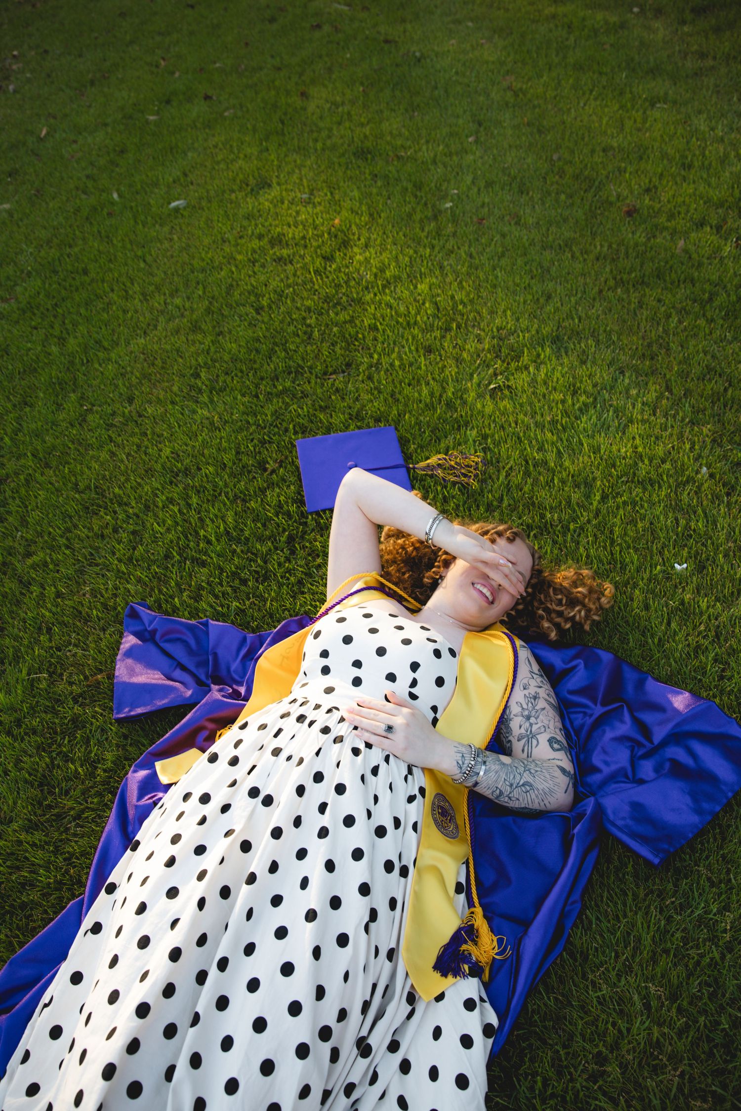 Graduate in polka dot dress lying on green grass with blue cap and yellow graduation stole.