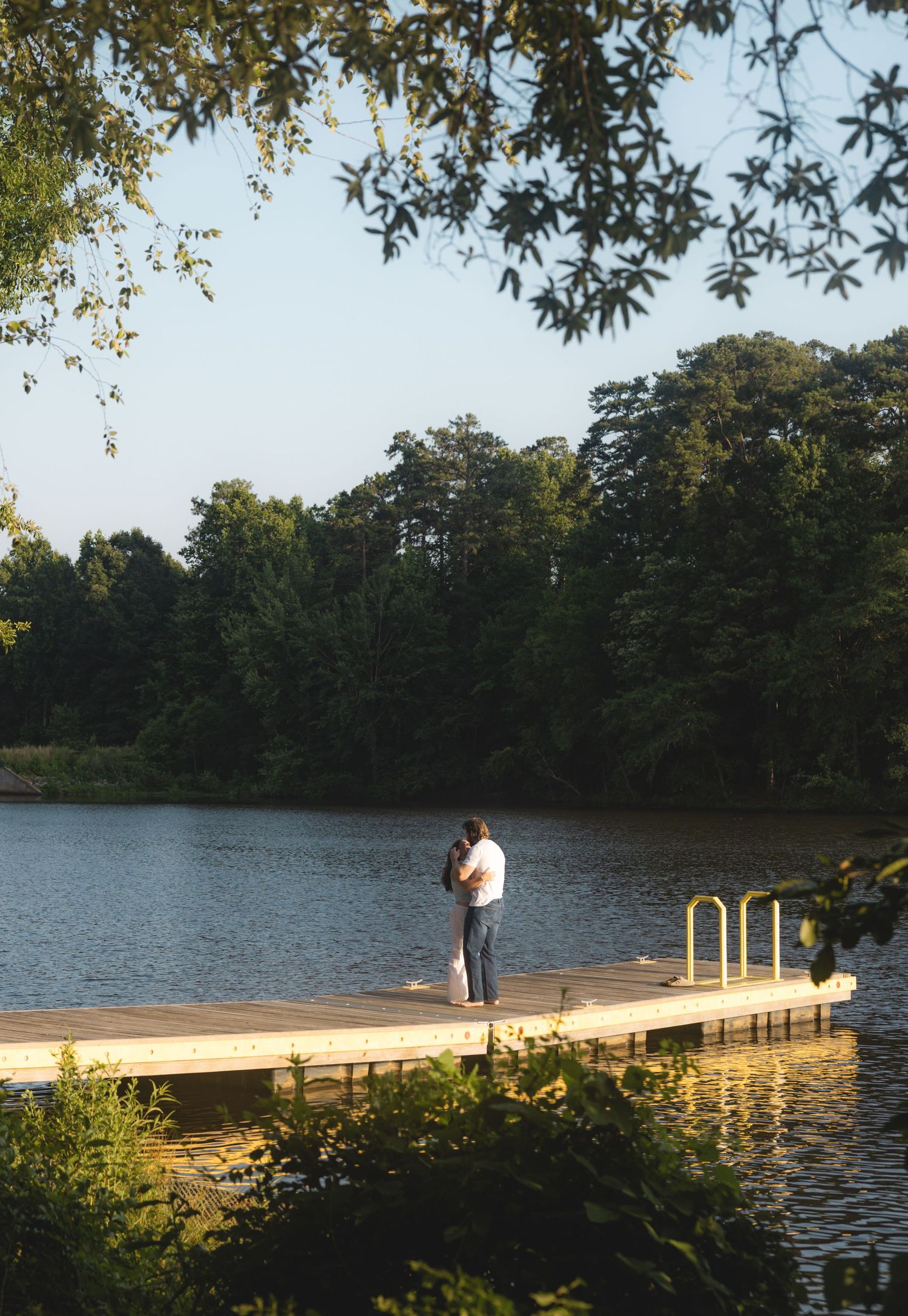 Couple embracing on a wooden dock overlooking a serene lake surrounded by lush green trees on a sunny day.