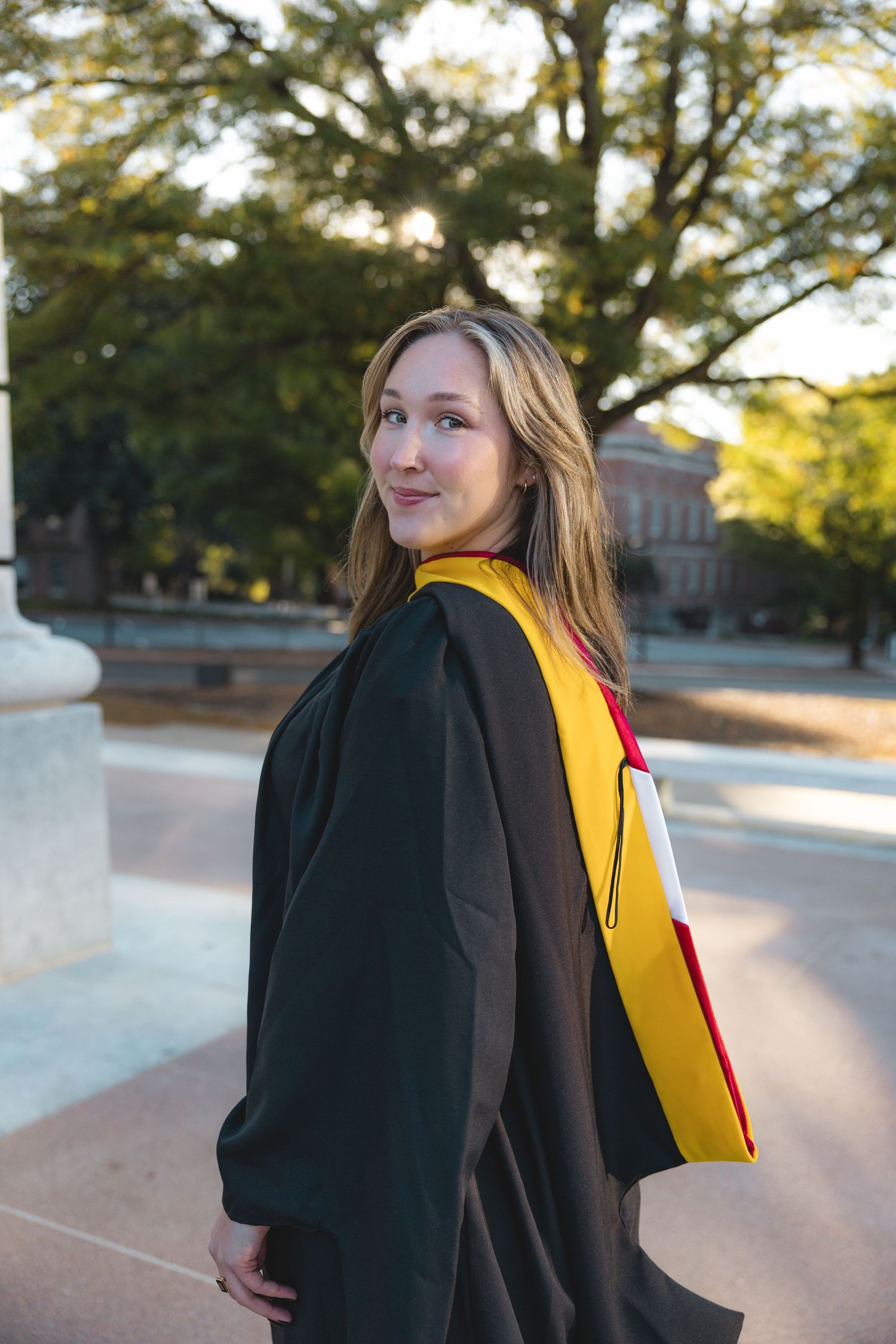 Young woman in black graduation gown with yellow and red sash smiles outdoors on a college campus.