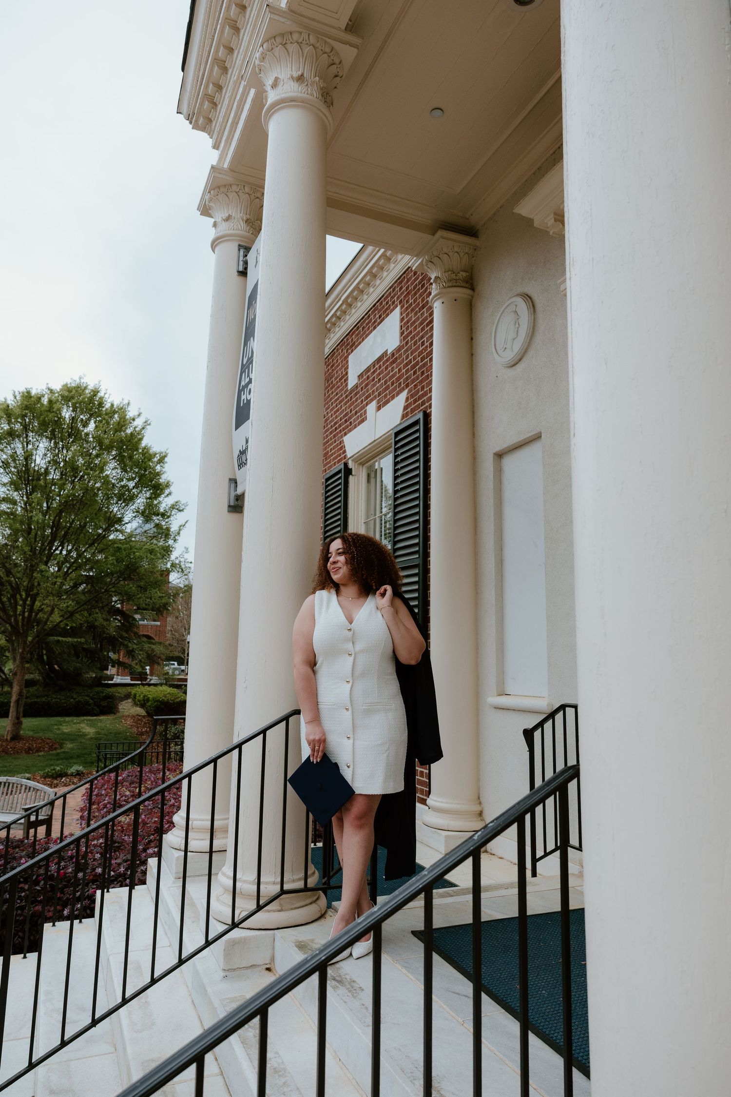 Woman in white dress holding graduation cap on steps of brick building with white columns.
