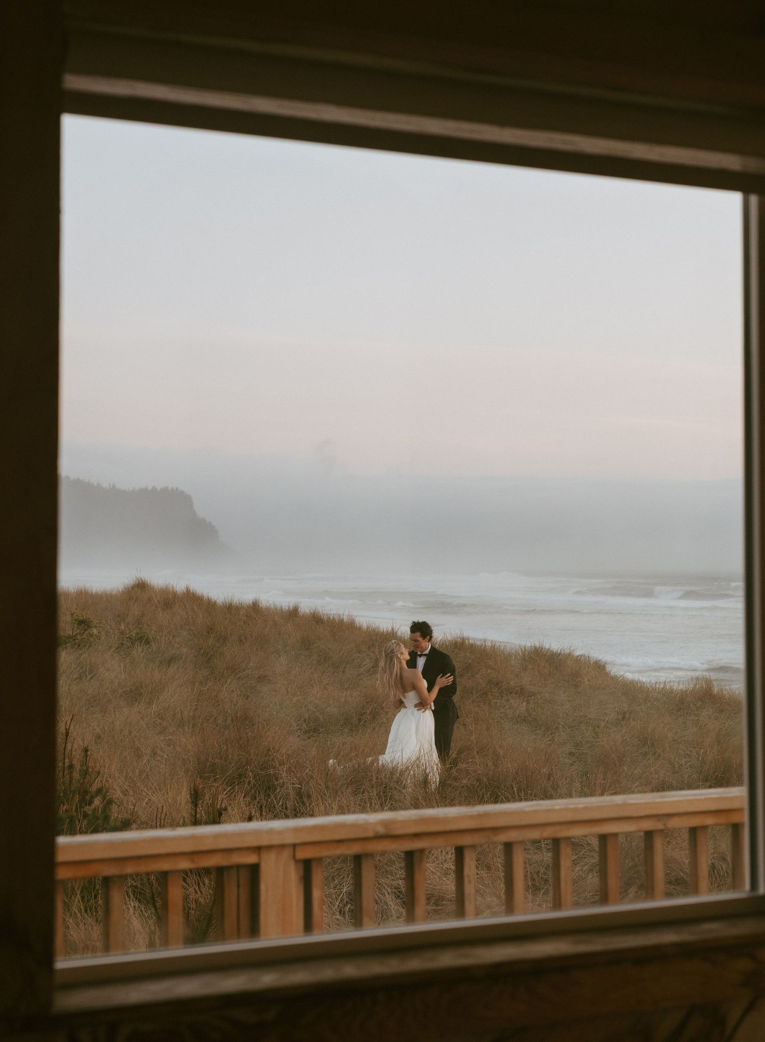 Couple embracing on coastal bluff at sunset, viewed through rustic window frame with ocean waves and misty cliffs behind them.