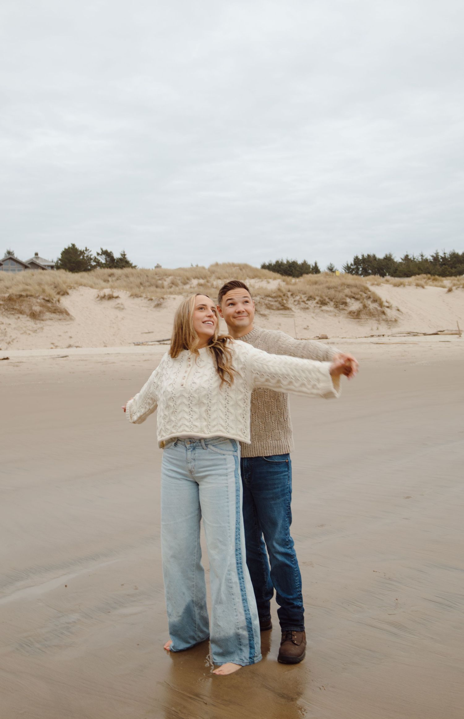 Happy couple embracing on a wide sandy beach with sand dunes and coastal vegetation behind them.