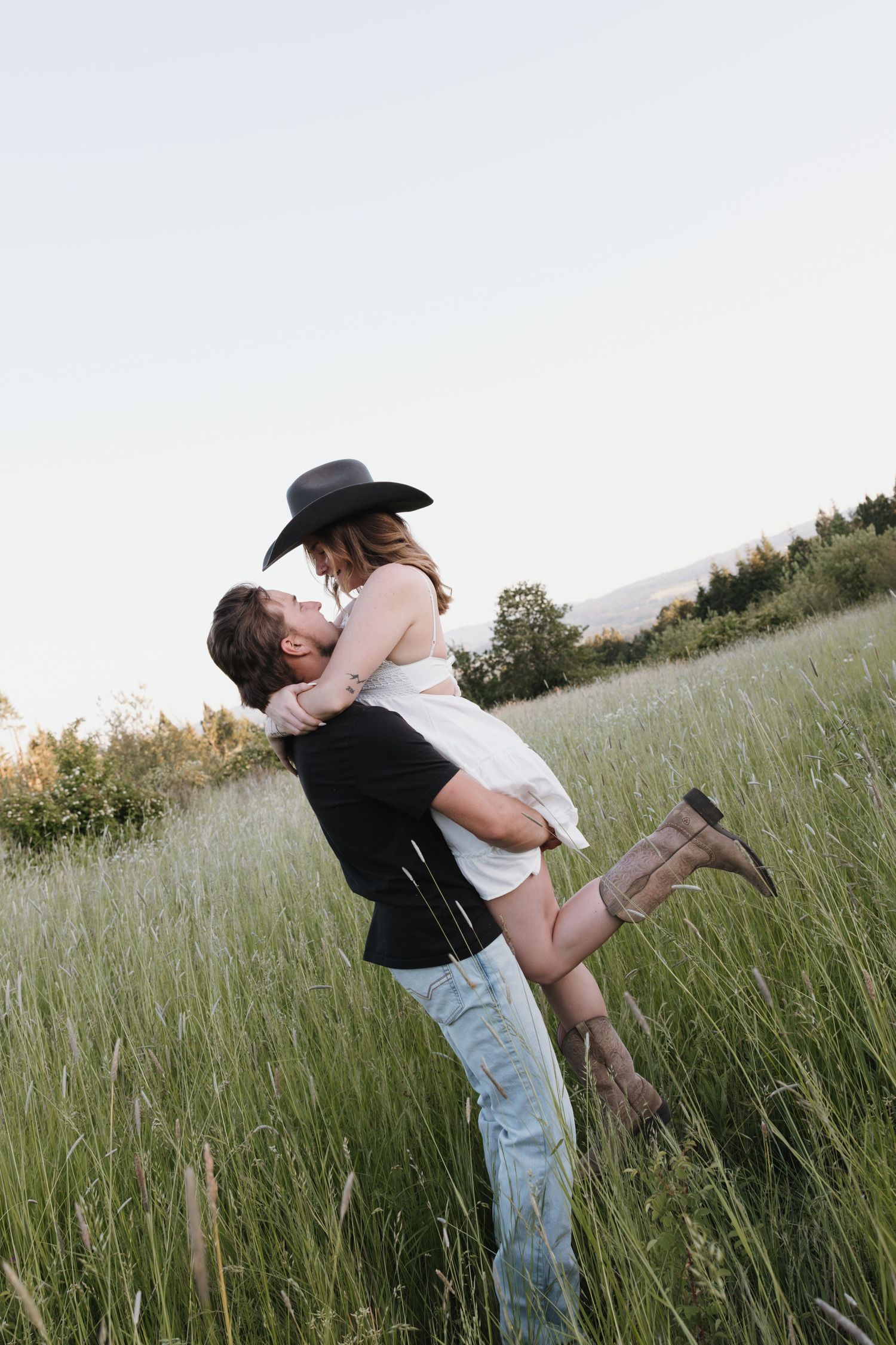 Couple sharing a romantic kiss in a lush green field, woman wearing a black cowboy hat and white dress lifted by her partner.