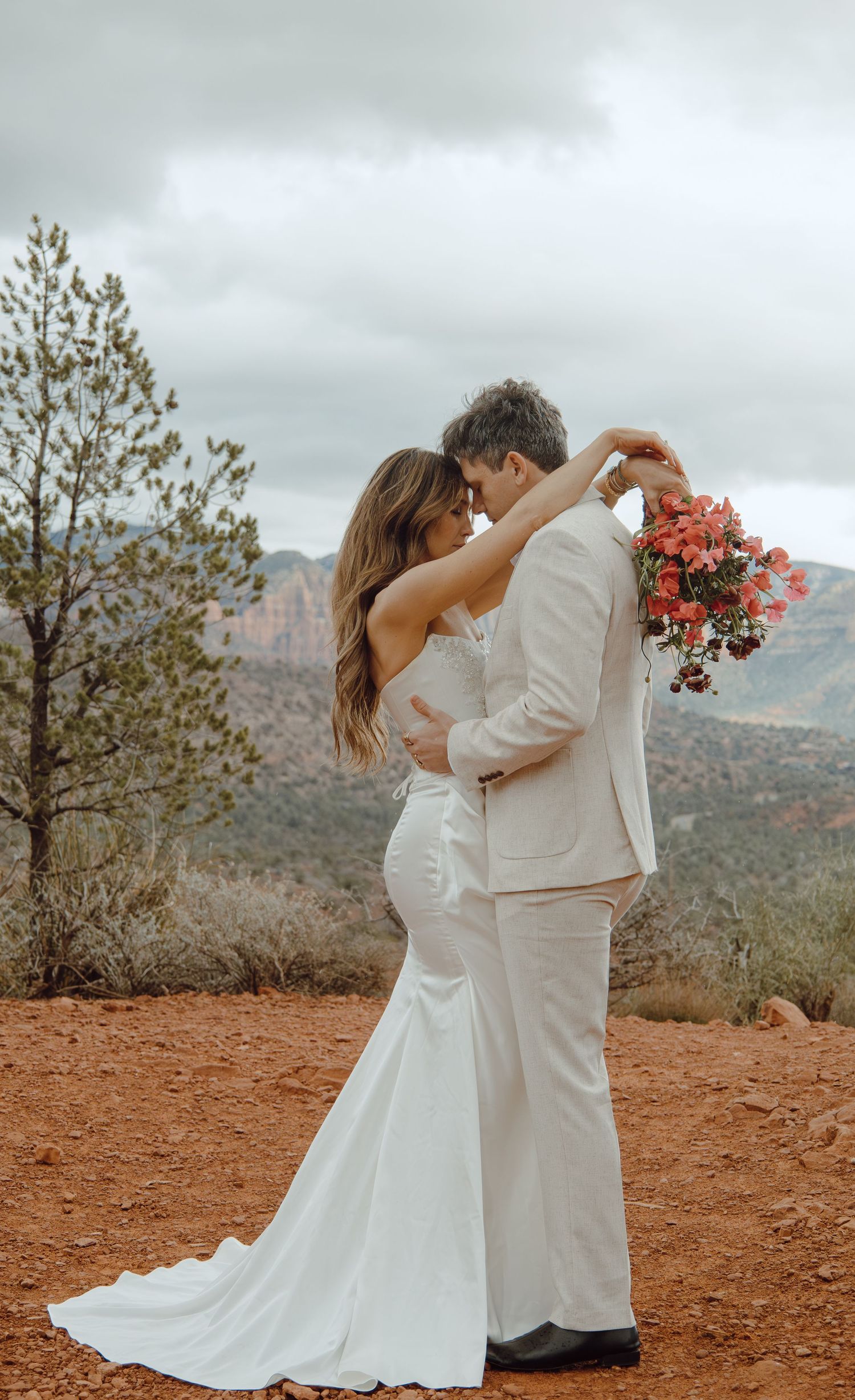 Wedding couple embracing with bride holding red bouquet against Sedona red rock scenery.
