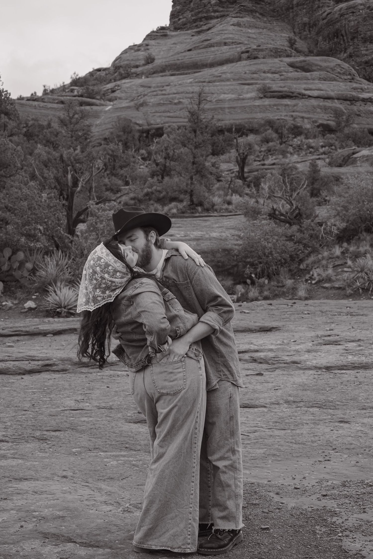 Black and white photo of a bearded man in cowboy hat kissing a woman against Sedona rock formations.