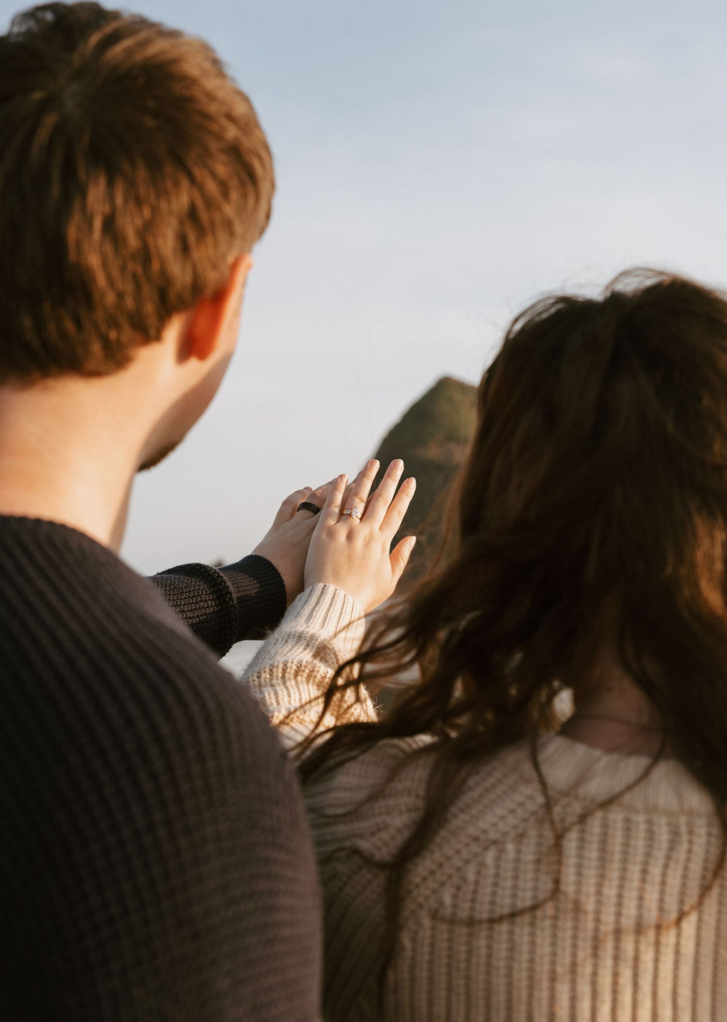 Couple holding hands outdoors, woman displaying engagement ring while wearing cream sweater with rocky landscape behind.