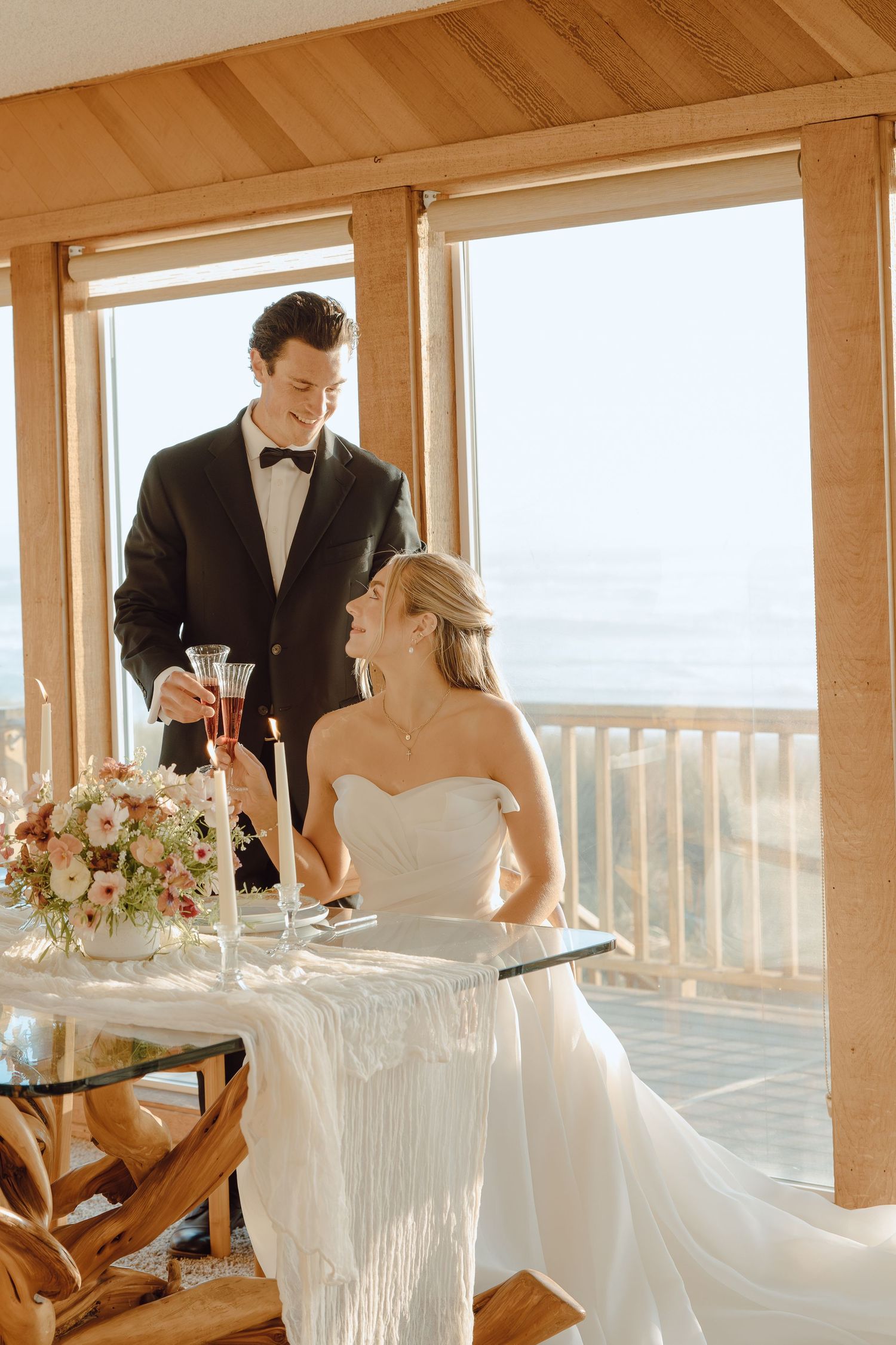Bride and groom toasting with champagne at elegant glass table with floral centerpiece.