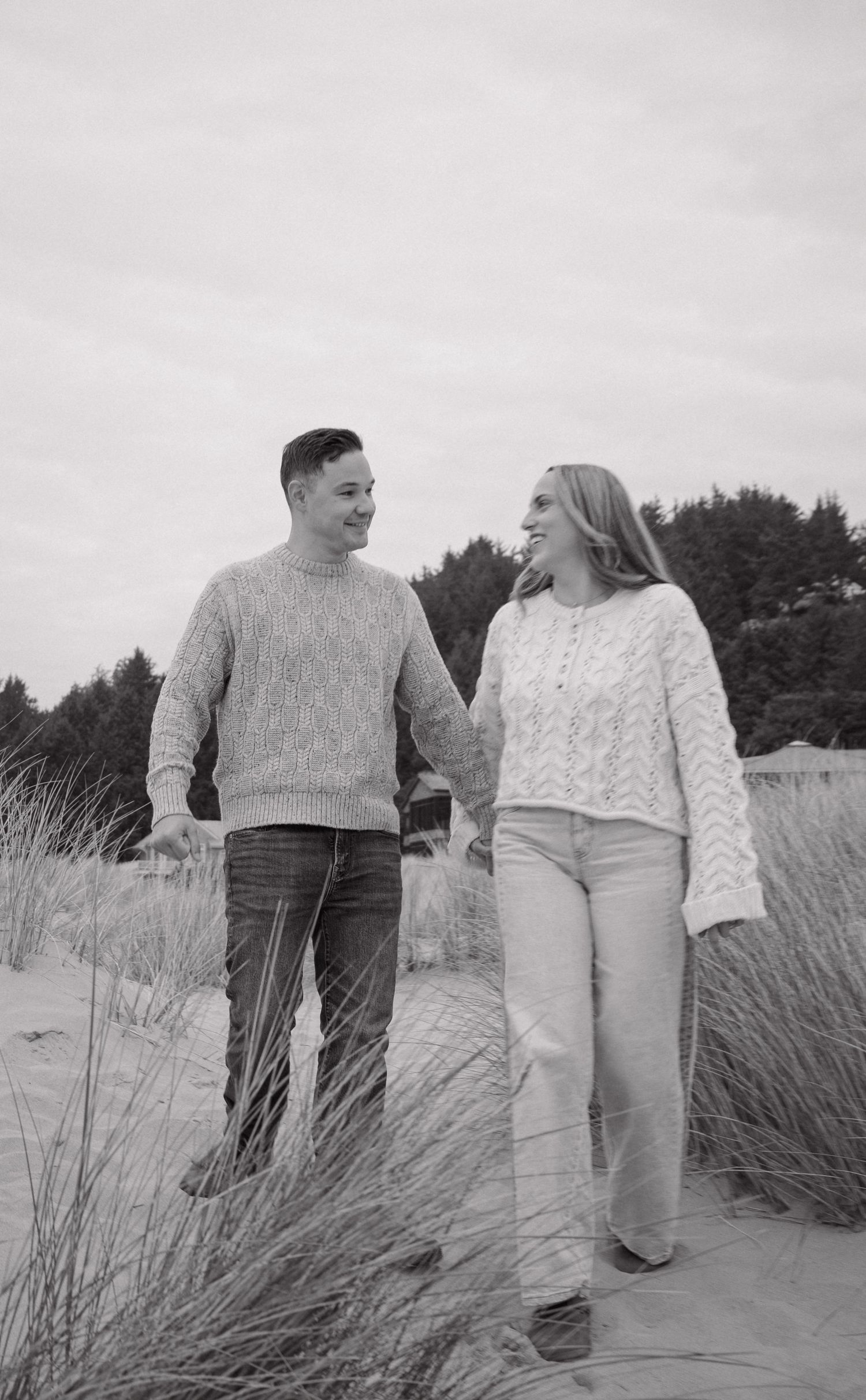 Black and white photo of couple holding hands and smiling while walking through beach grass dunes.