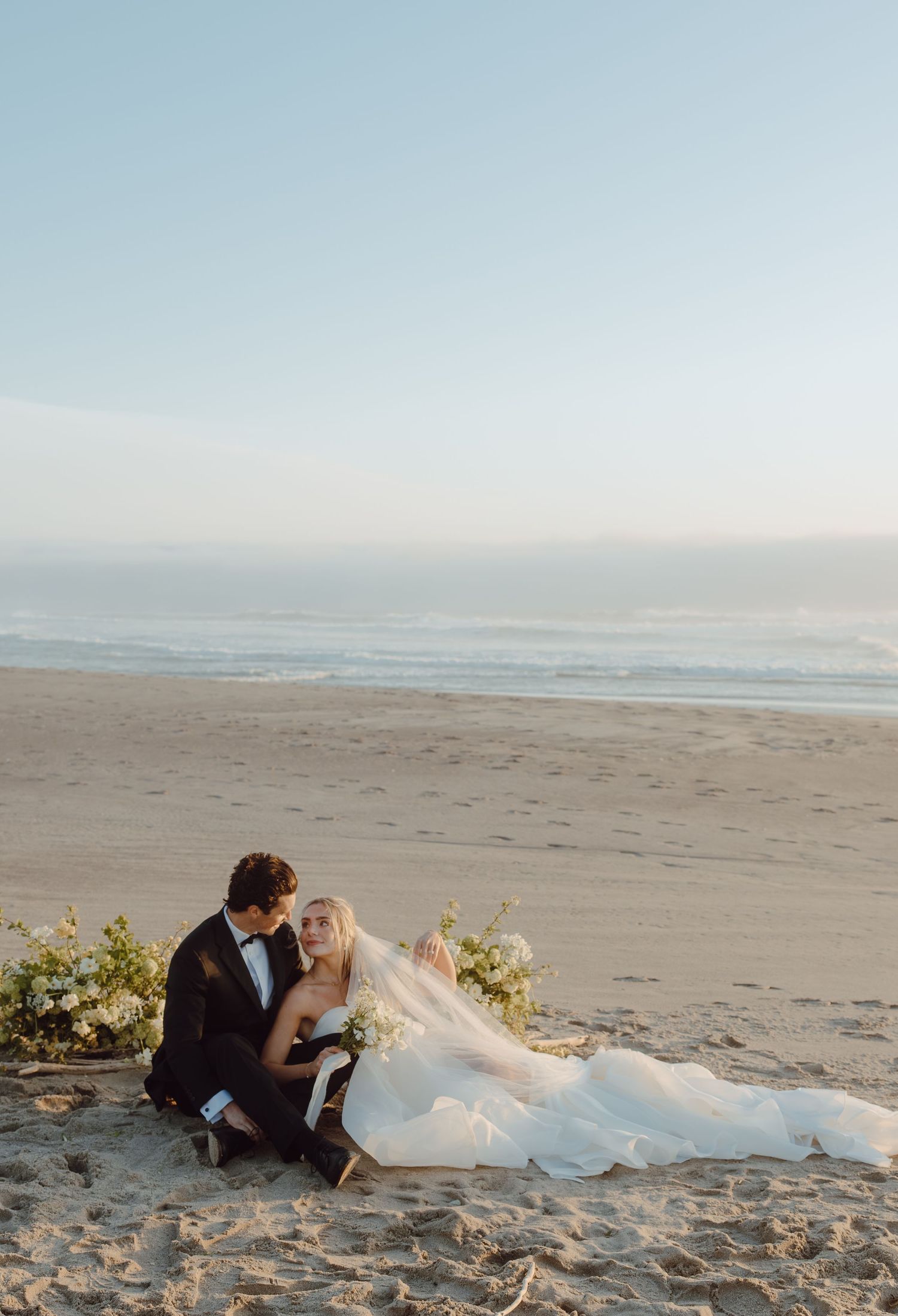Bride and groom sitting together on sandy beach at sunset, with ocean waves in the background, bride in flowing white gown.