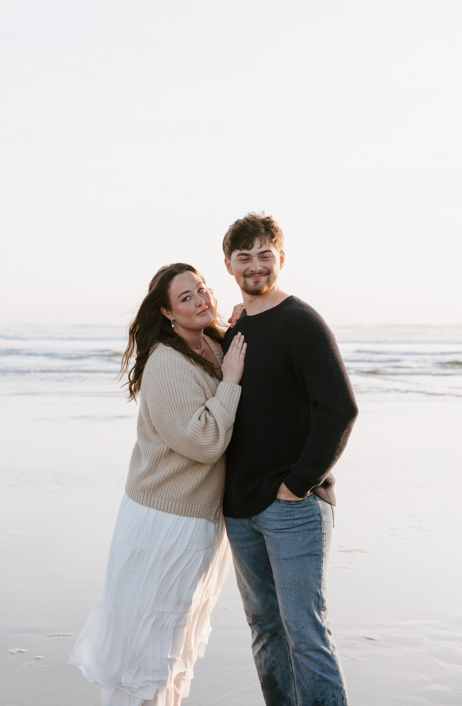 Couple embracing on a beach at sunset, woman in beige sweater and white skirt, man in black sweater and jeans.