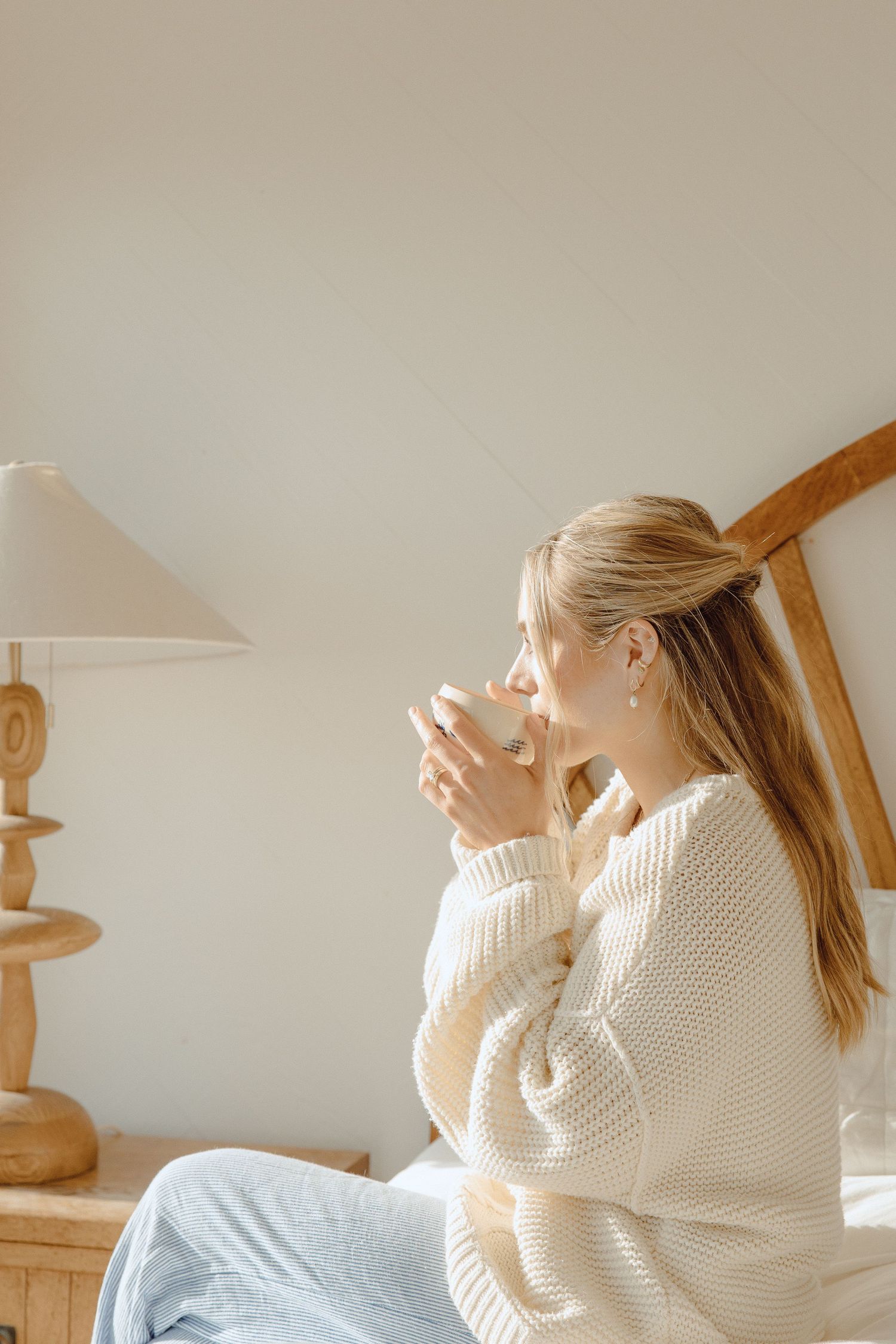 Young blonde woman in cream knit sweater sitting on bed sipping hot drink in bright cozy bedroom.