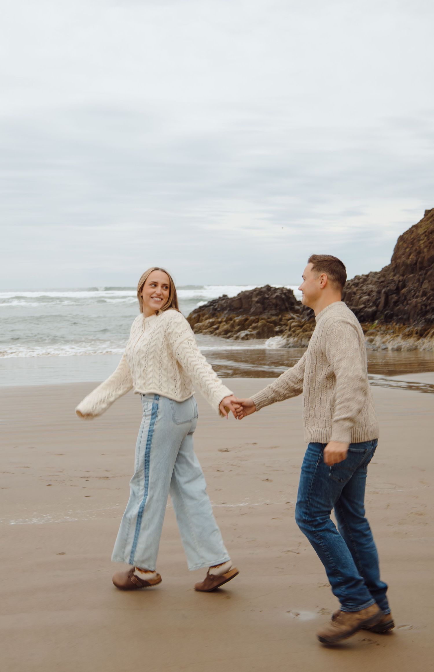 Couple holding hands walking on a sandy beach with rocky cliffs and ocean waves in the background.