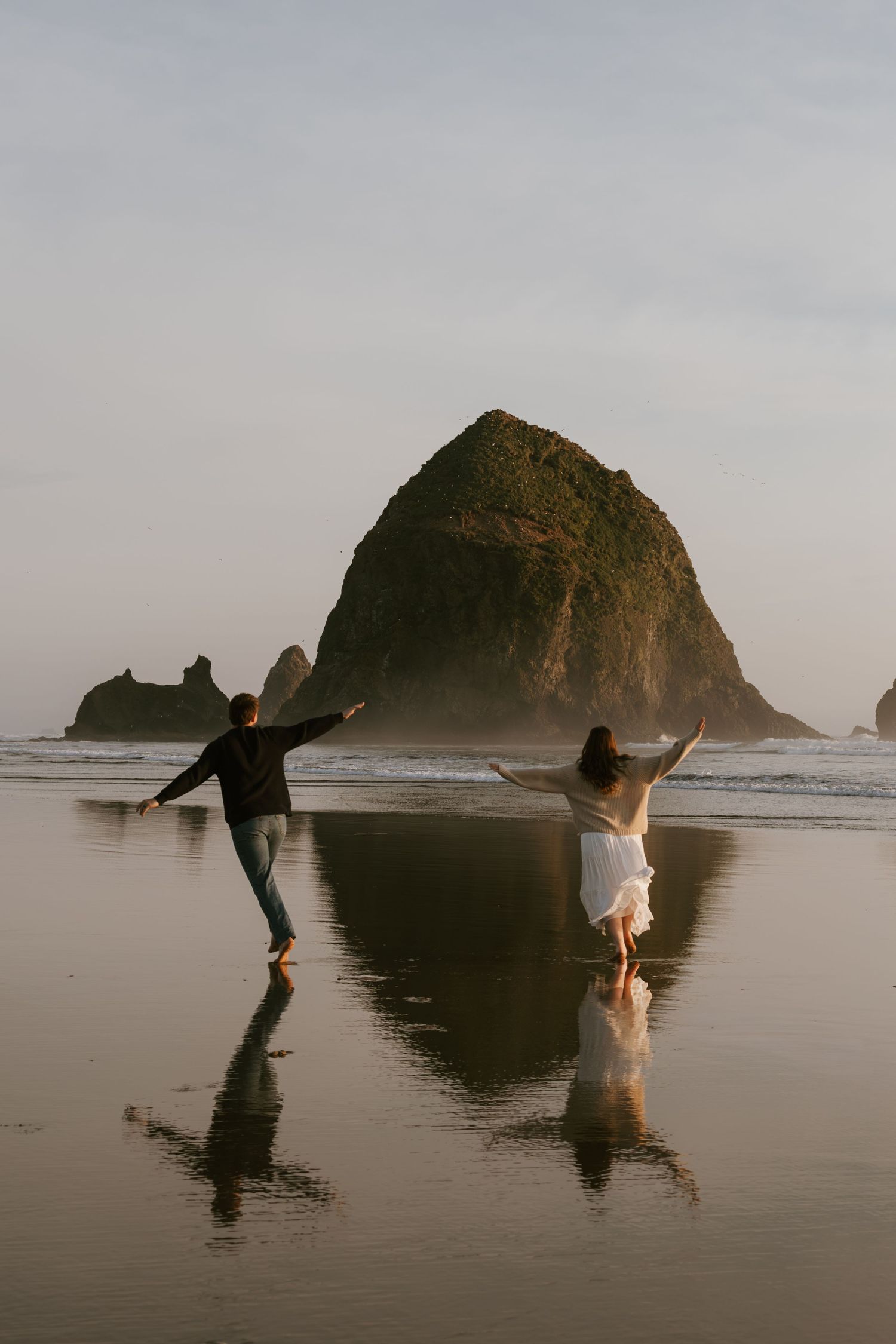 Couple dancing joyfully on reflective beach sand with iconic Haystack Rock at Cannon Beach, Oregon in the background.
