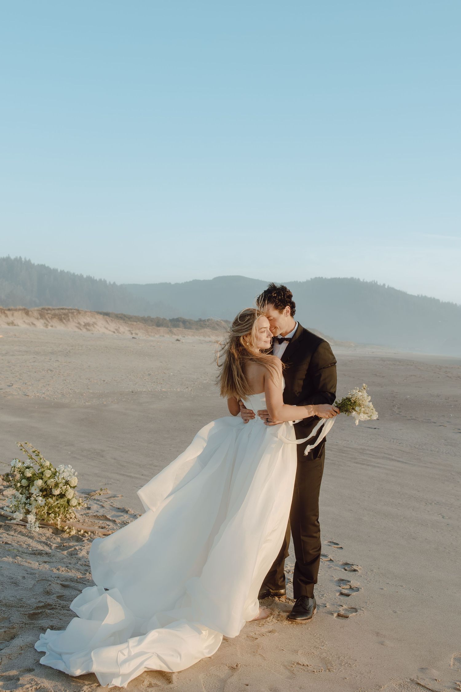 Bride and groom share romantic kiss on sandy beach, bride in flowing white gown holding bouquet, mountains in background.