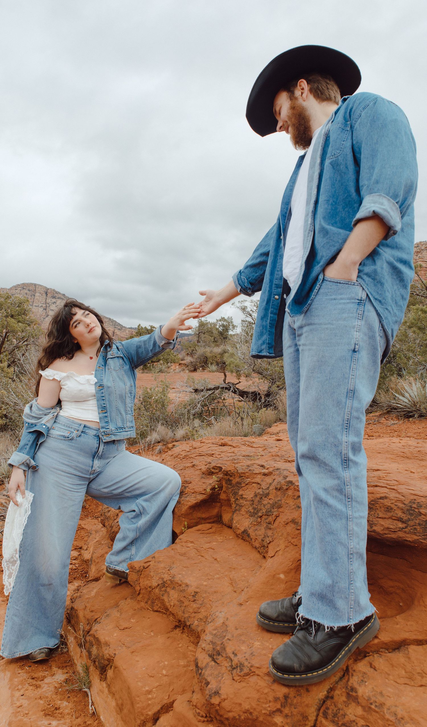 Man in cowboy hat reaching hand to woman sitting on red sandstone rocks in Sedona, Arizona.