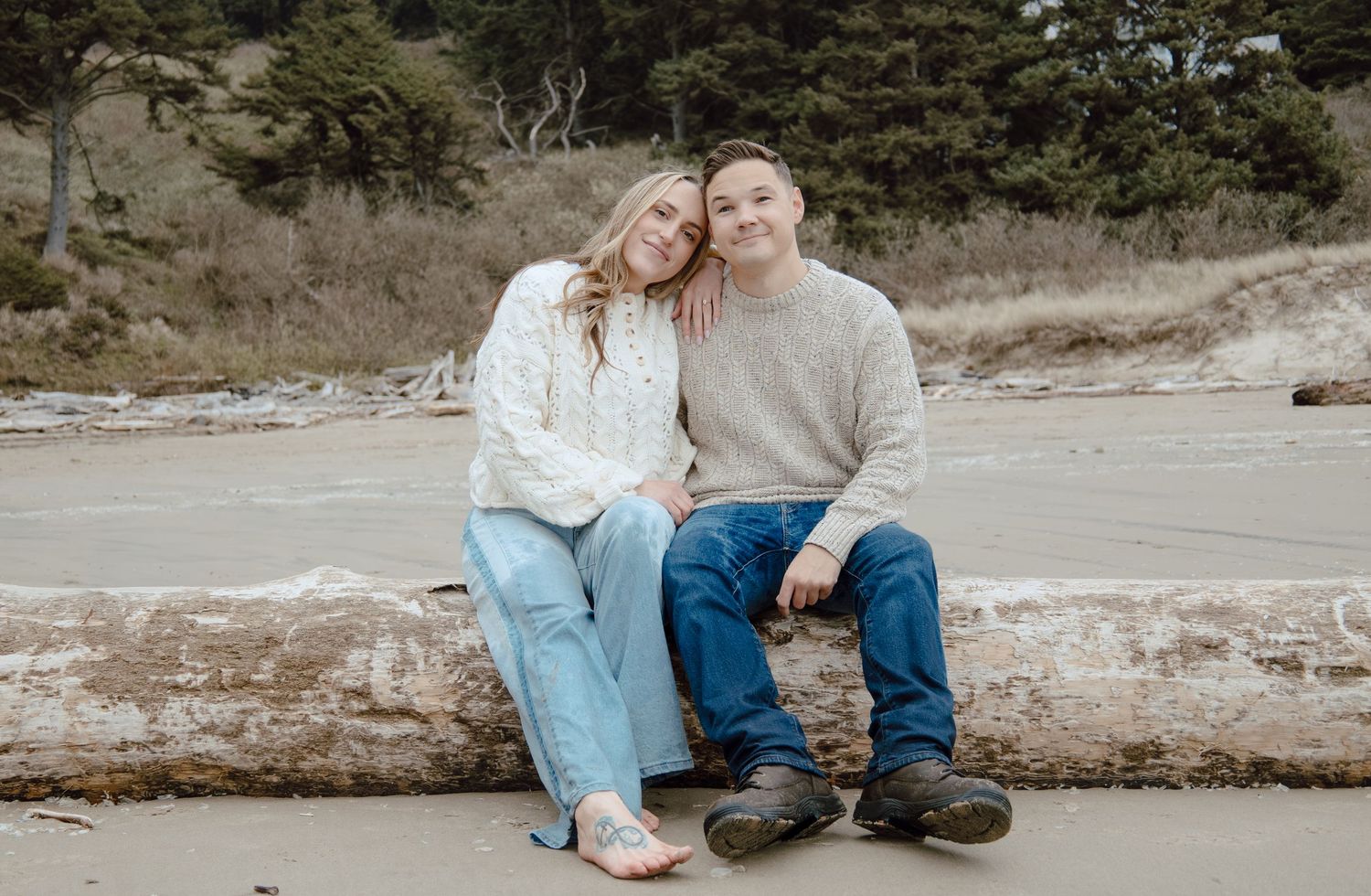 Couple sitting closely together on driftwood at a beach, smiling warmly at each other.