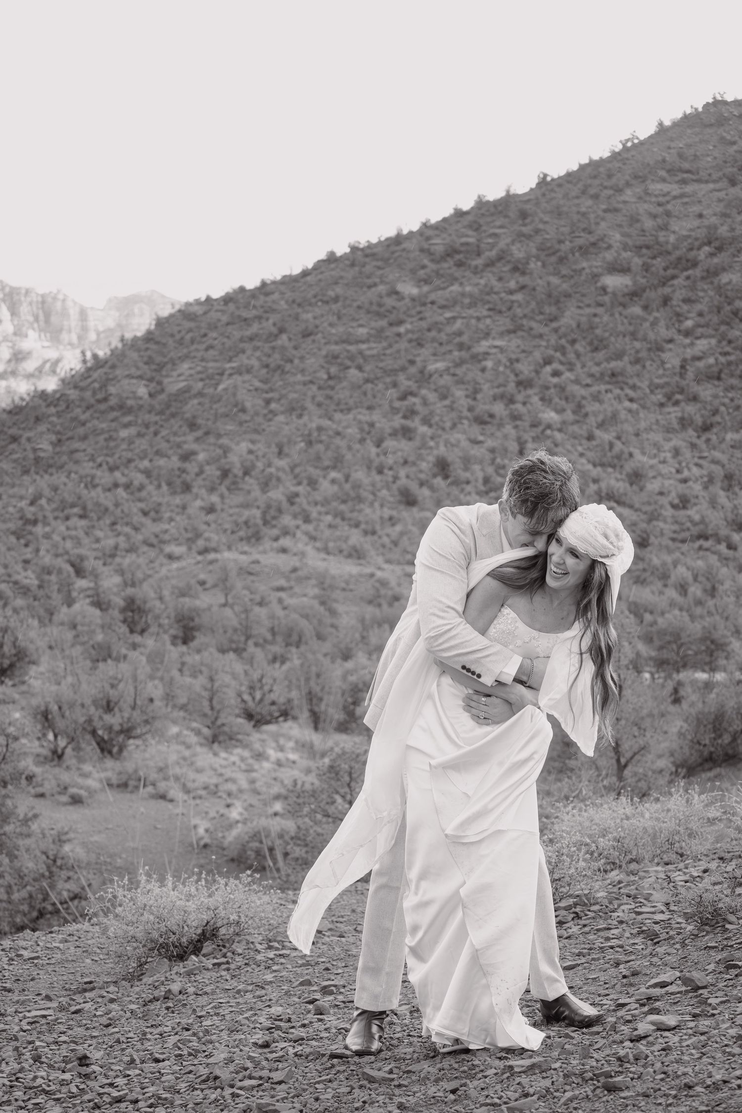 Black and white photo of groom dipping bride on rocky terrain with mountain backdrop.