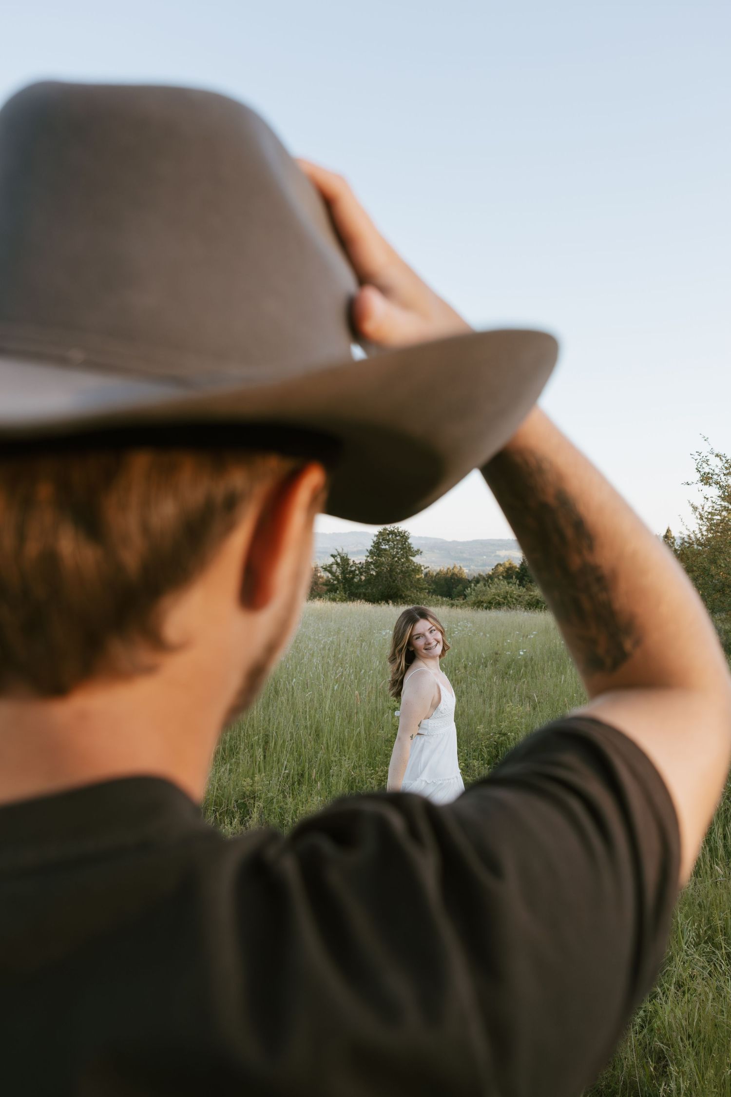 Man in black shirt and cowboy hat looking at smiling woman in white dress standing in a green field.