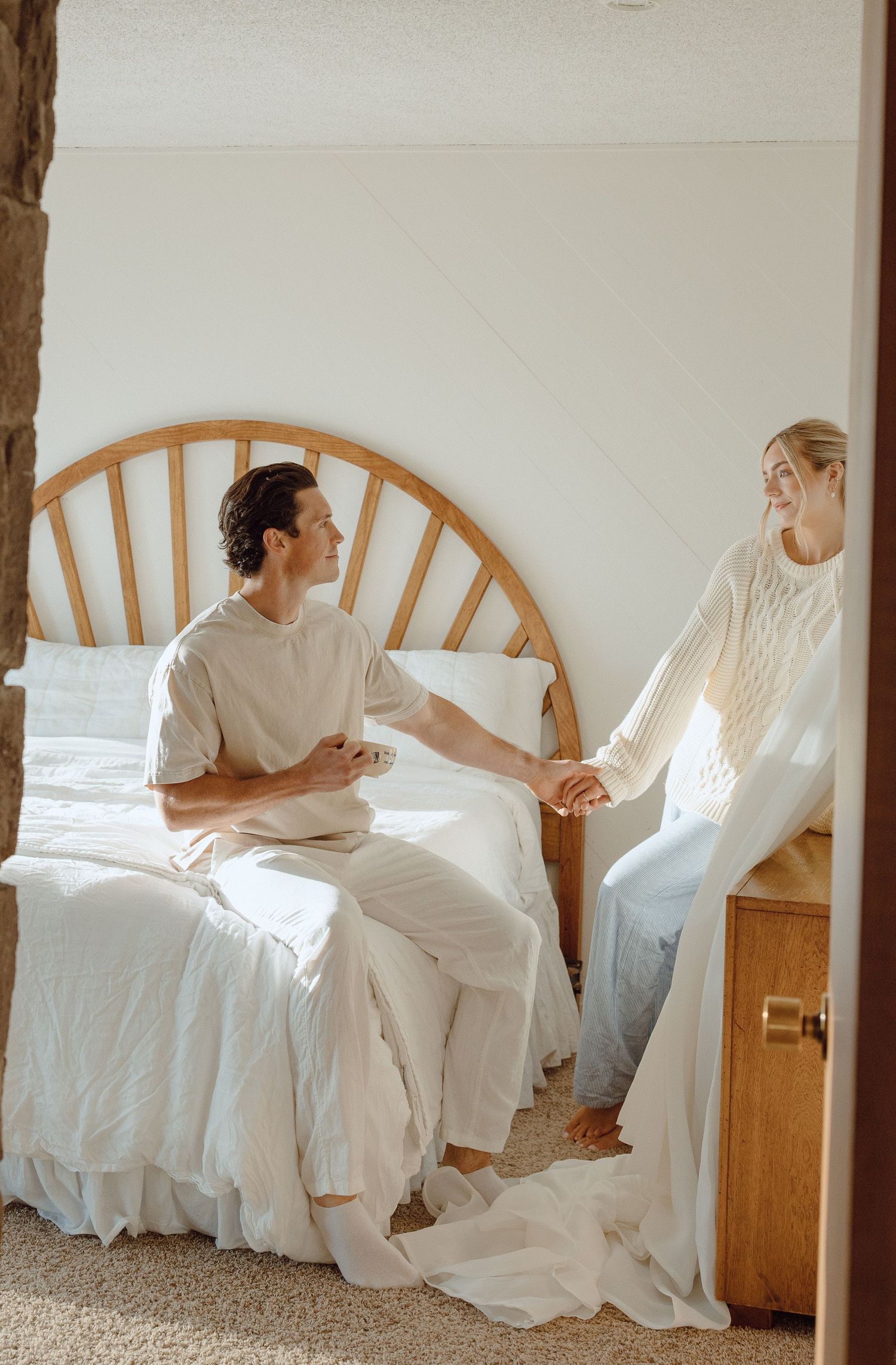 Couple in white outfits holding hands on a sunlit bed with wooden headboard.