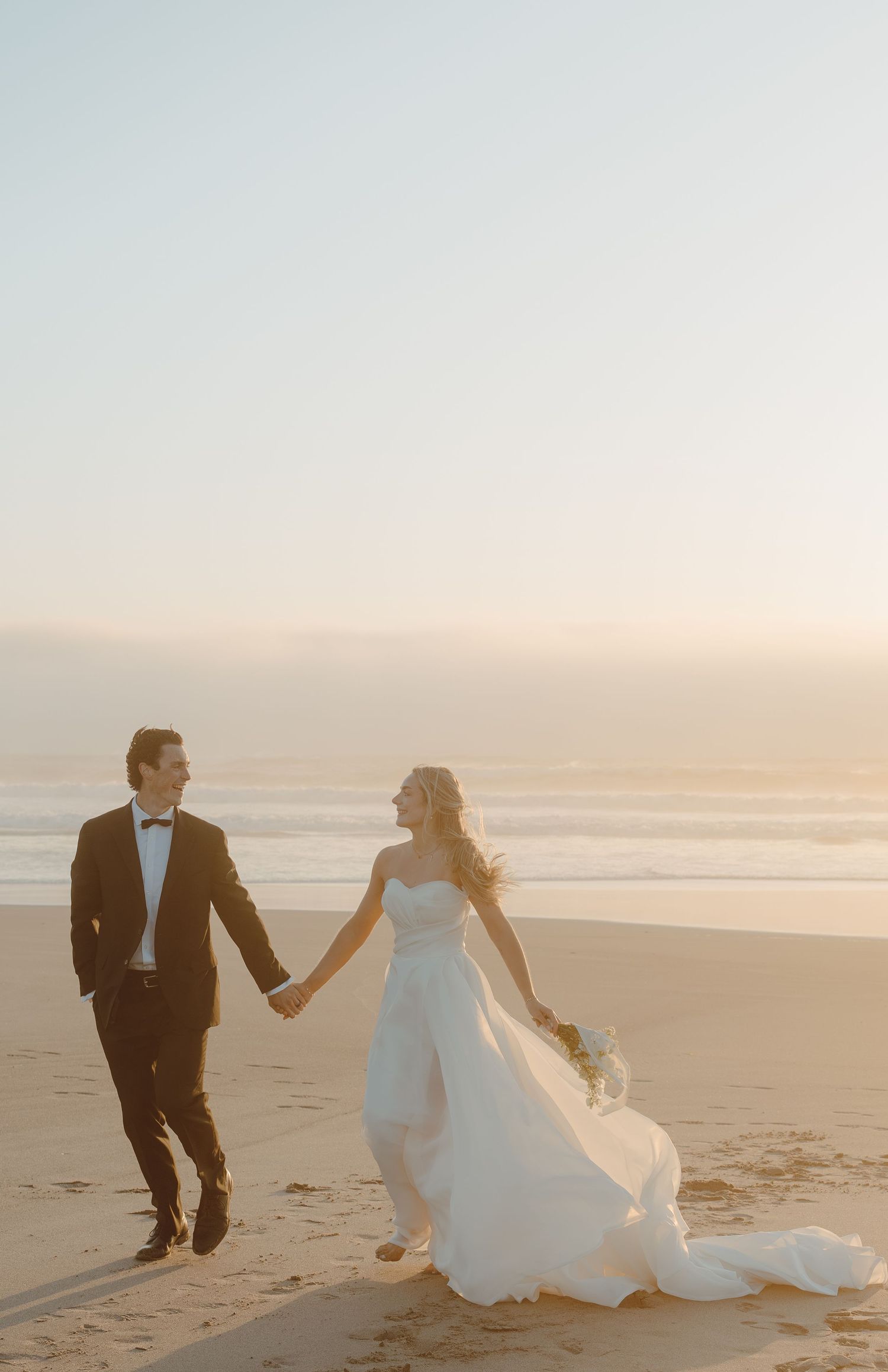 Bride and groom holding hands walking on beach at golden sunset.