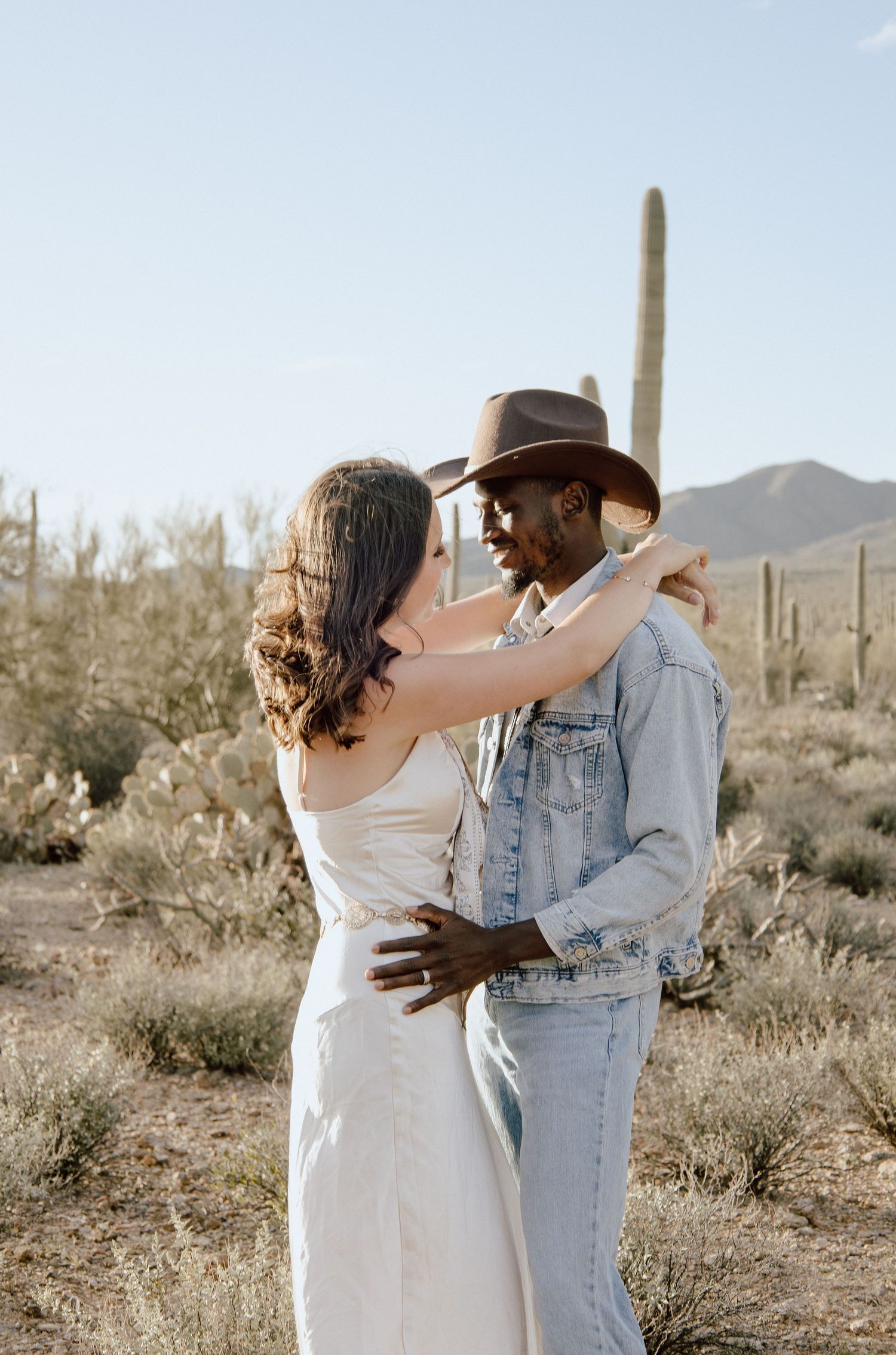 Couple sharing romantic moment in Sonoran Desert, woman adjusting mans brown cowboy hat surrounded by cacti.
