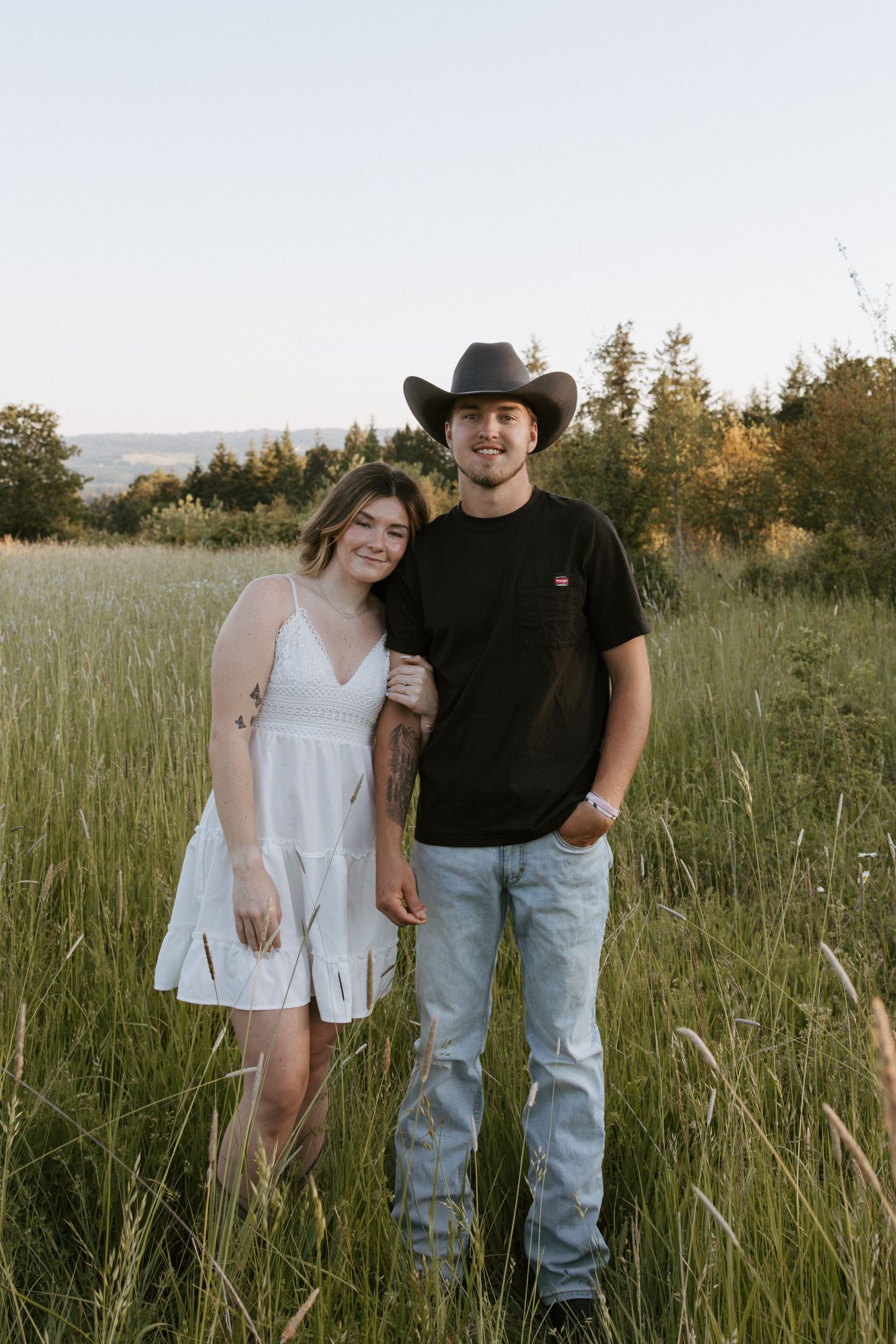 A couple poses in a grassy field, woman in white dress, man in black tee, jeans, and cowboy hat.