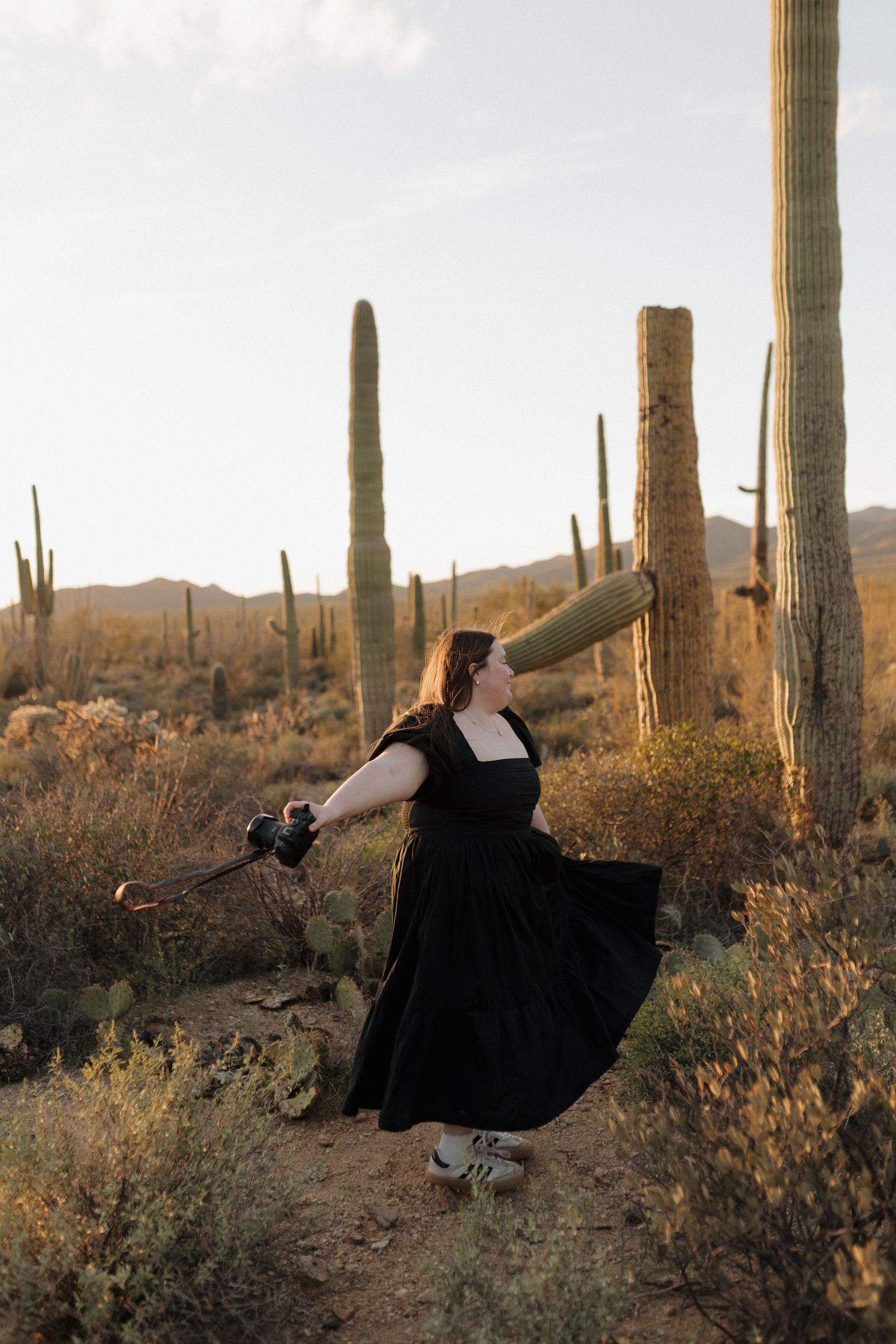 Woman in black dress twirling among tall saguaro cacti in Arizona desert at sunset.