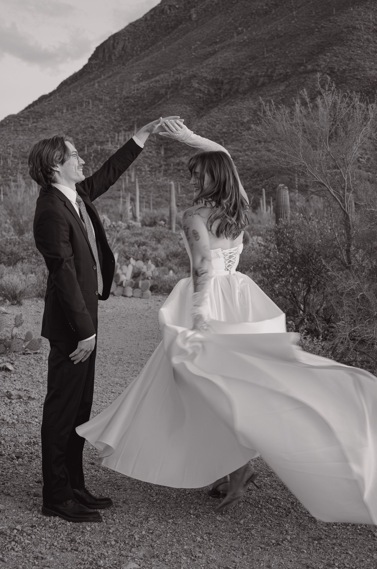 Black and white photo of groom spinning bride in wedding dress on desert path with cacti backdrop.