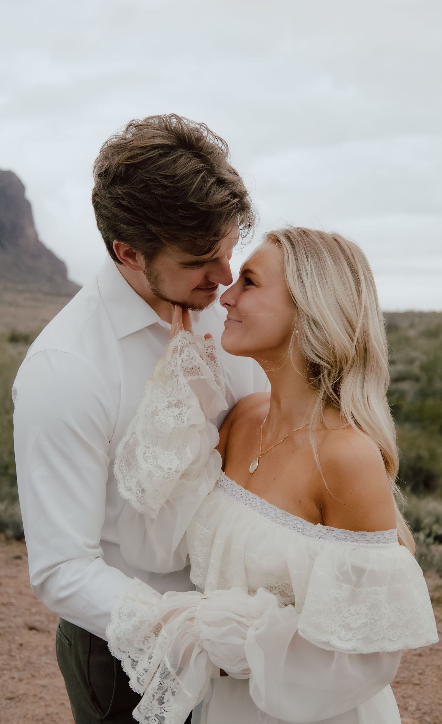 Smiling couple nearly kisses outdoors, woman in off-shoulder lace dress, red rock formations behind.