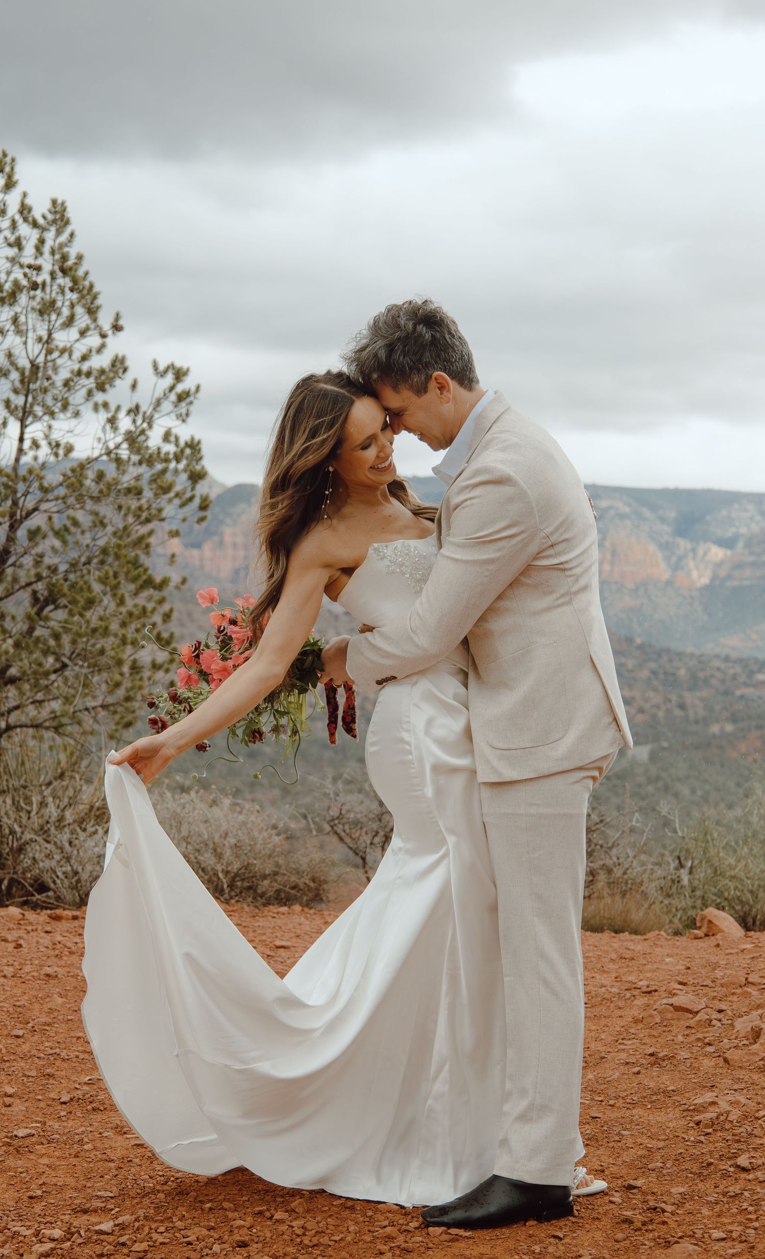 Bride in white gown holds bouquet while groom in tan suit embraces her in Sedona.