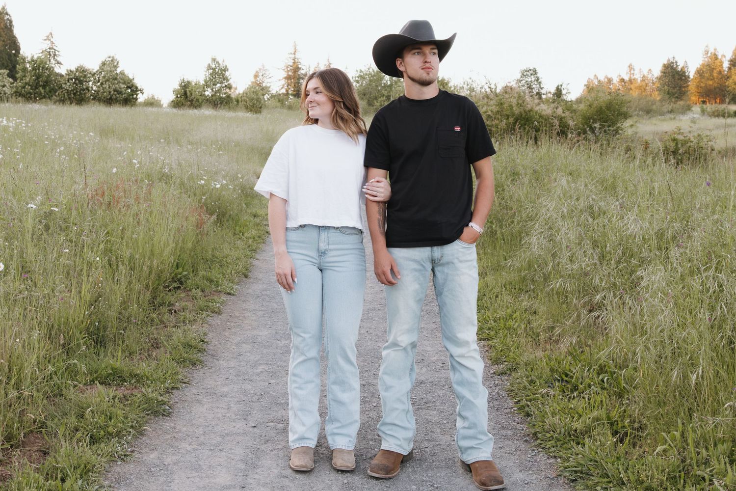 Young couple standing on dirt path in field, woman in white tee and jeans, man in black tee and cowboy hat.