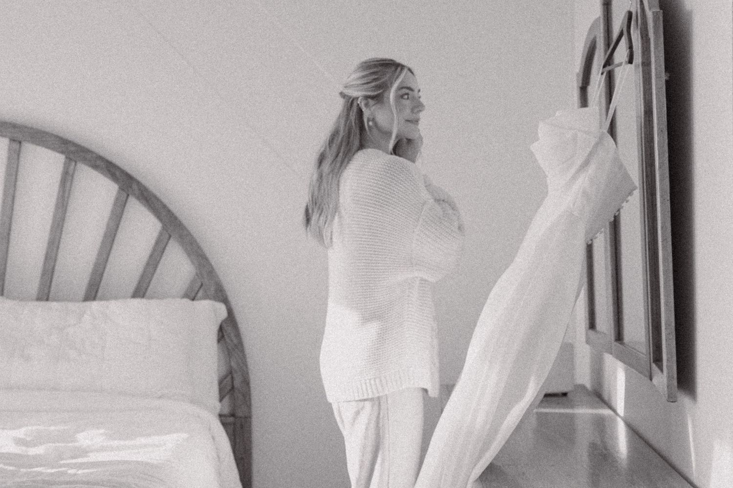 Woman in white sweater standing by a bed holding sheer curtains in a black and white bedroom photo.
