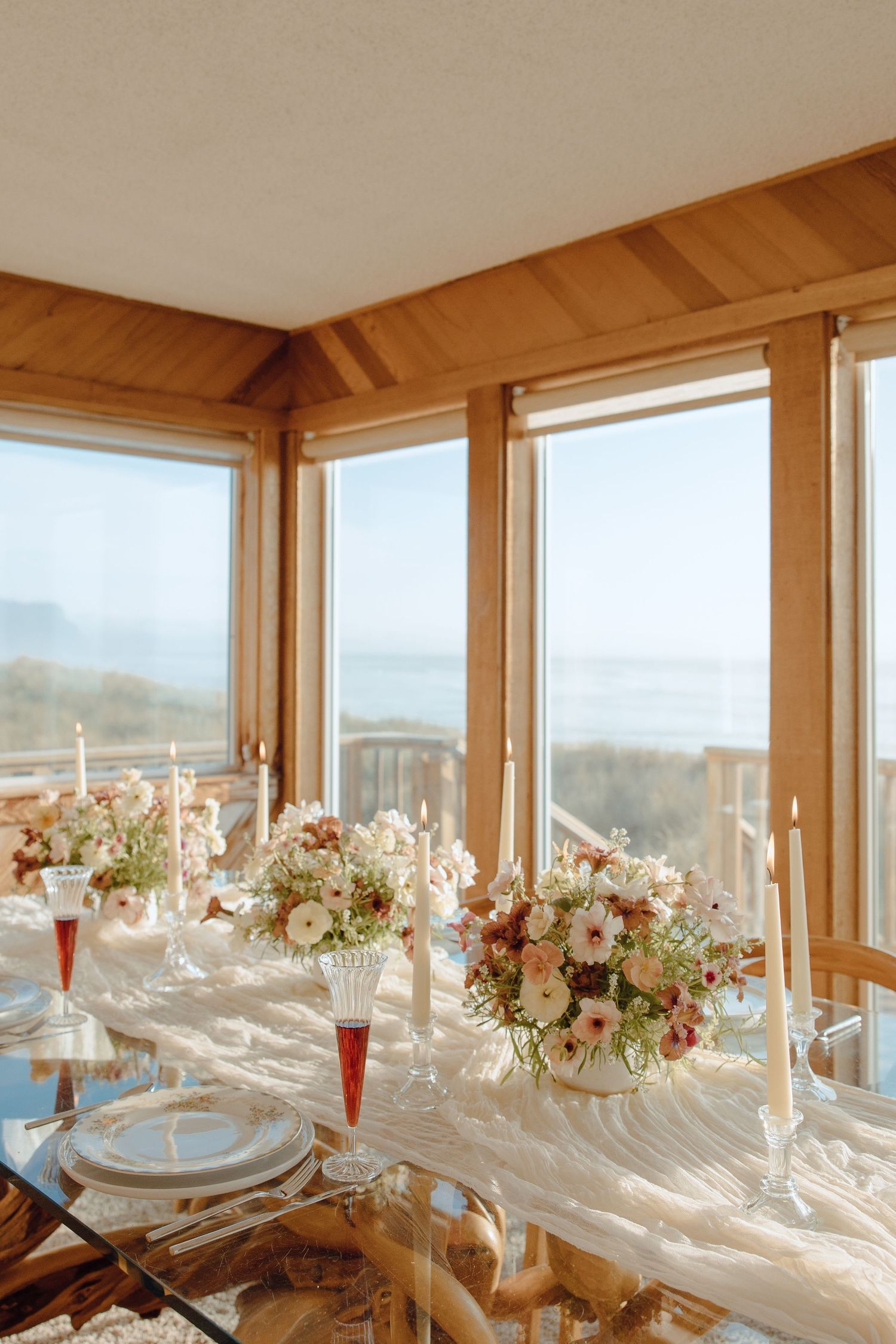 Elegant coastal wedding tablescape with floral centerpieces, taper candles, and ocean views through wood-framed windows.
