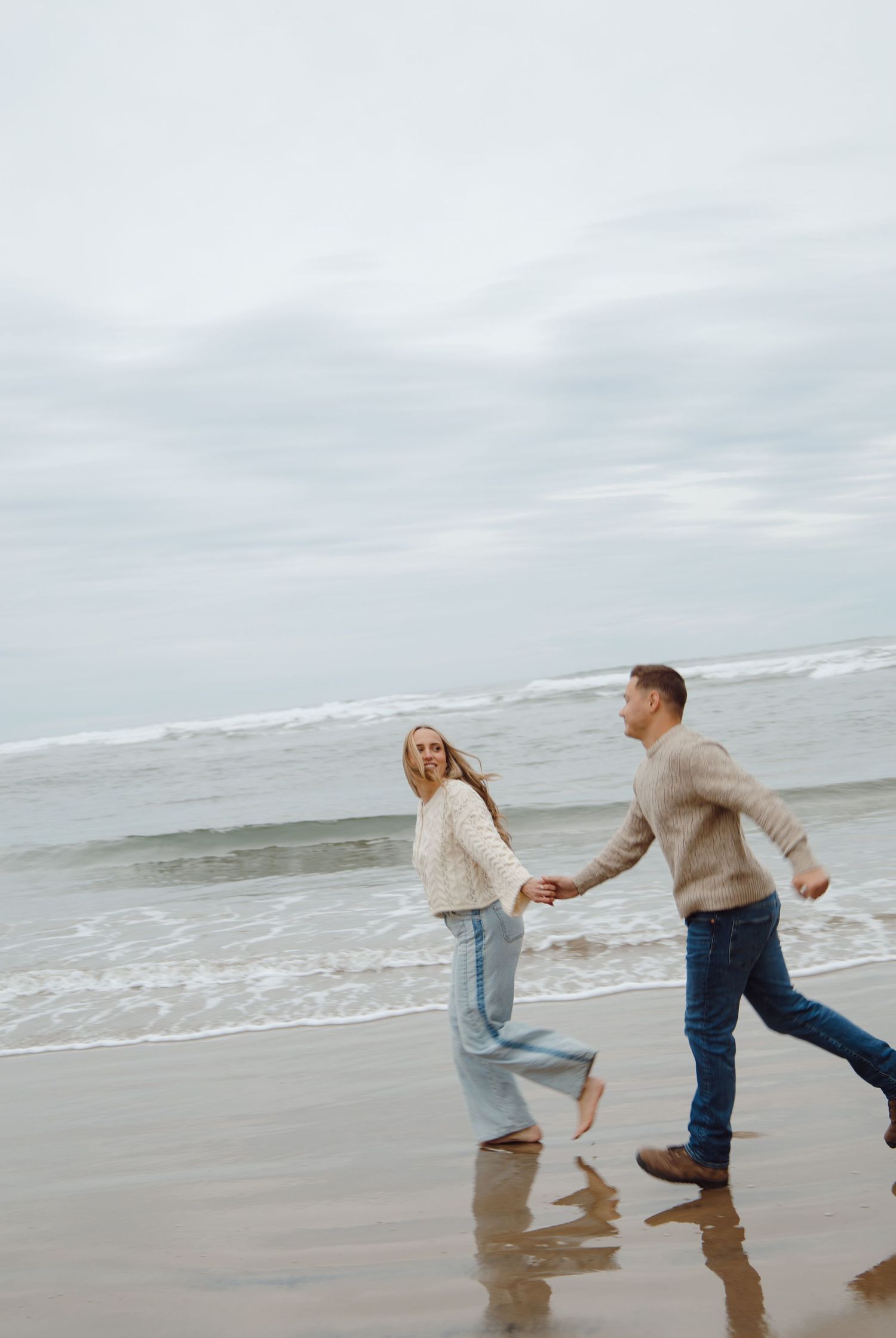 Couple in matching knit sweaters running hand-in-hand along a cloudy ocean beach shoreline.