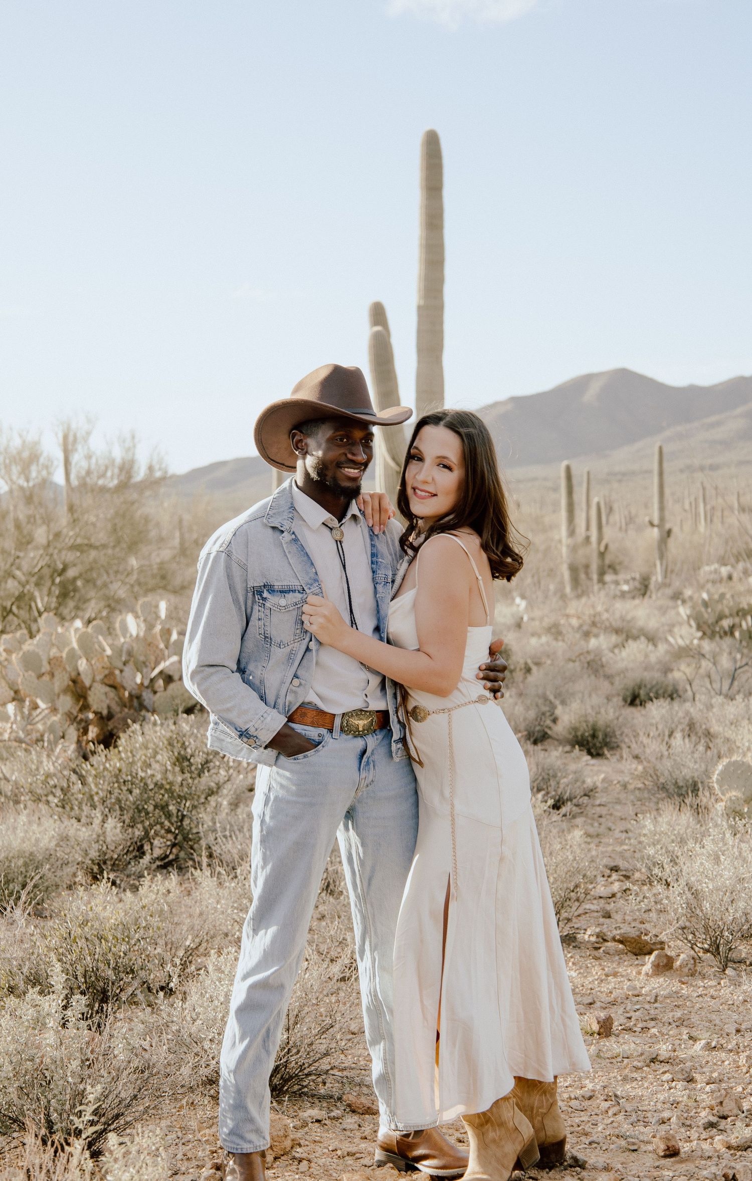 Interracial couple posing in Sonoran Desert, man in cowboy hat and denim, woman in white midi dress with cowboy boots.