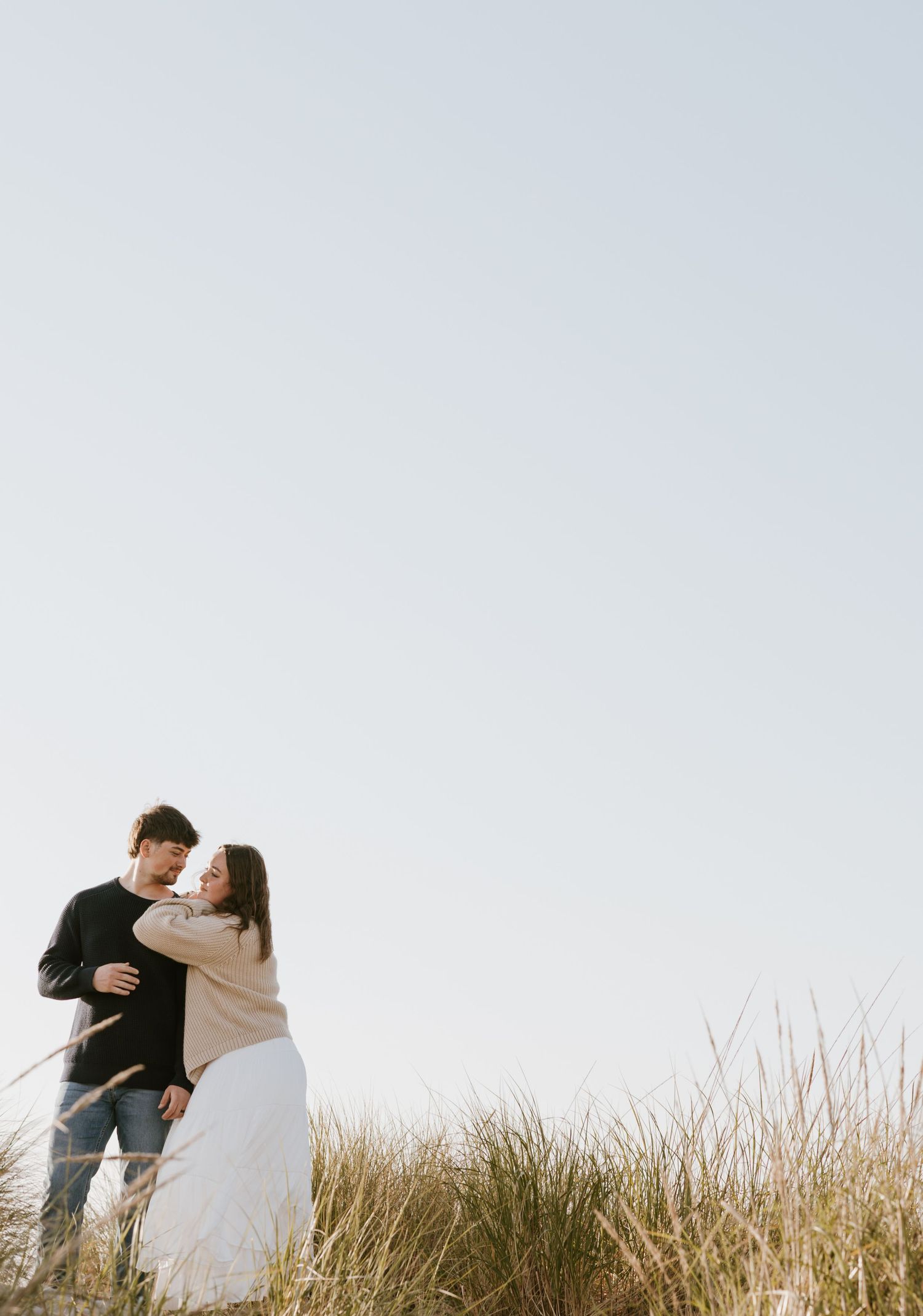 Couple embracing romantically among tall grass dunes under a clear sky in a serene outdoor setting.