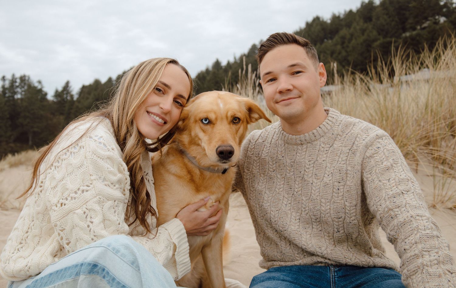 Smiling couple posing with their golden retriever dog among sand dunes with evergreen trees behind them.