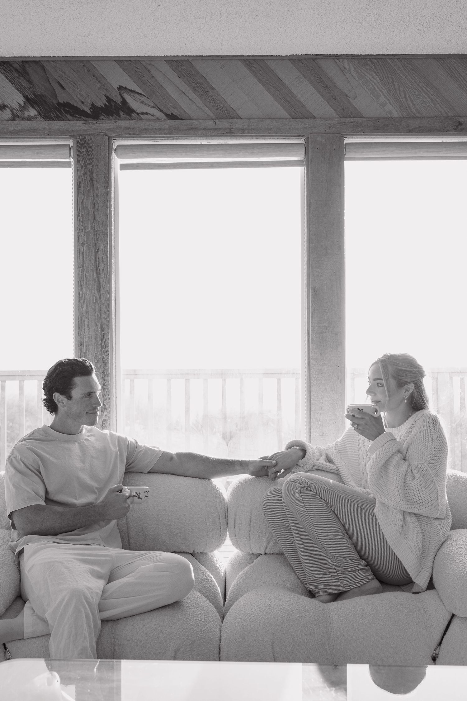 Black and white photo of a couple relaxing on a sofa by large windows, sharing a peaceful moment with coffee.