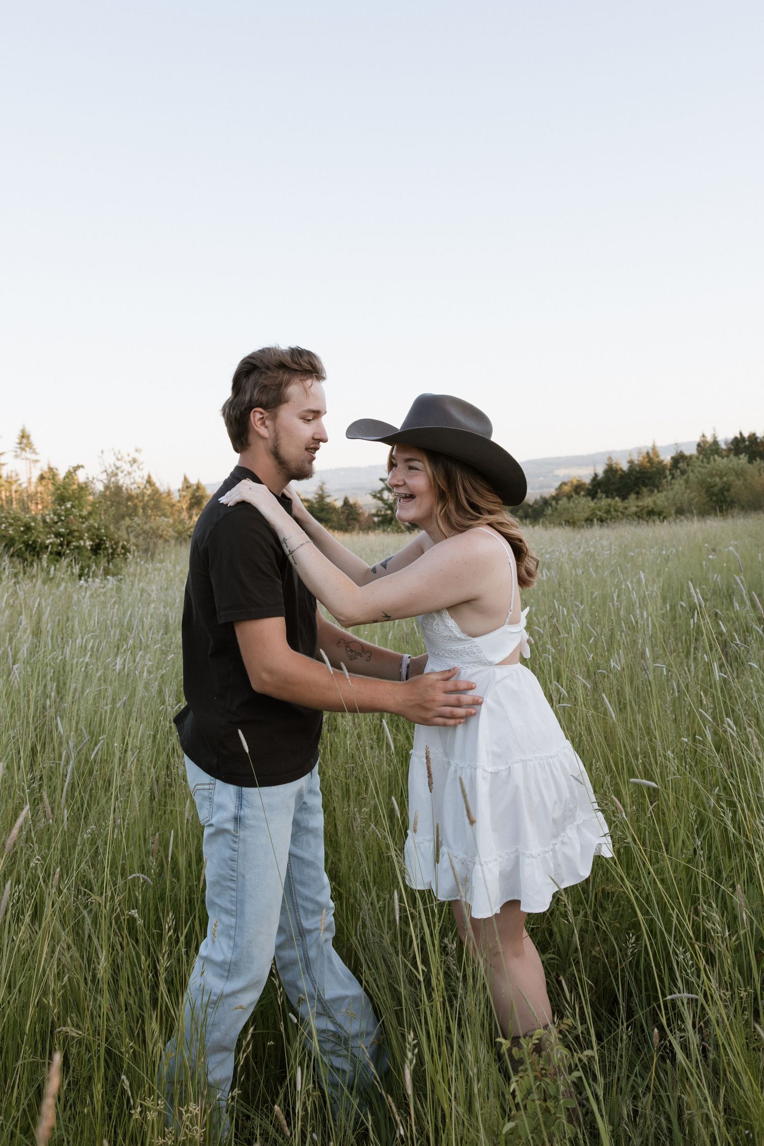 A couple dances in a lush green field, woman in white dress and cowboy hat, man in black shirt and jeans.